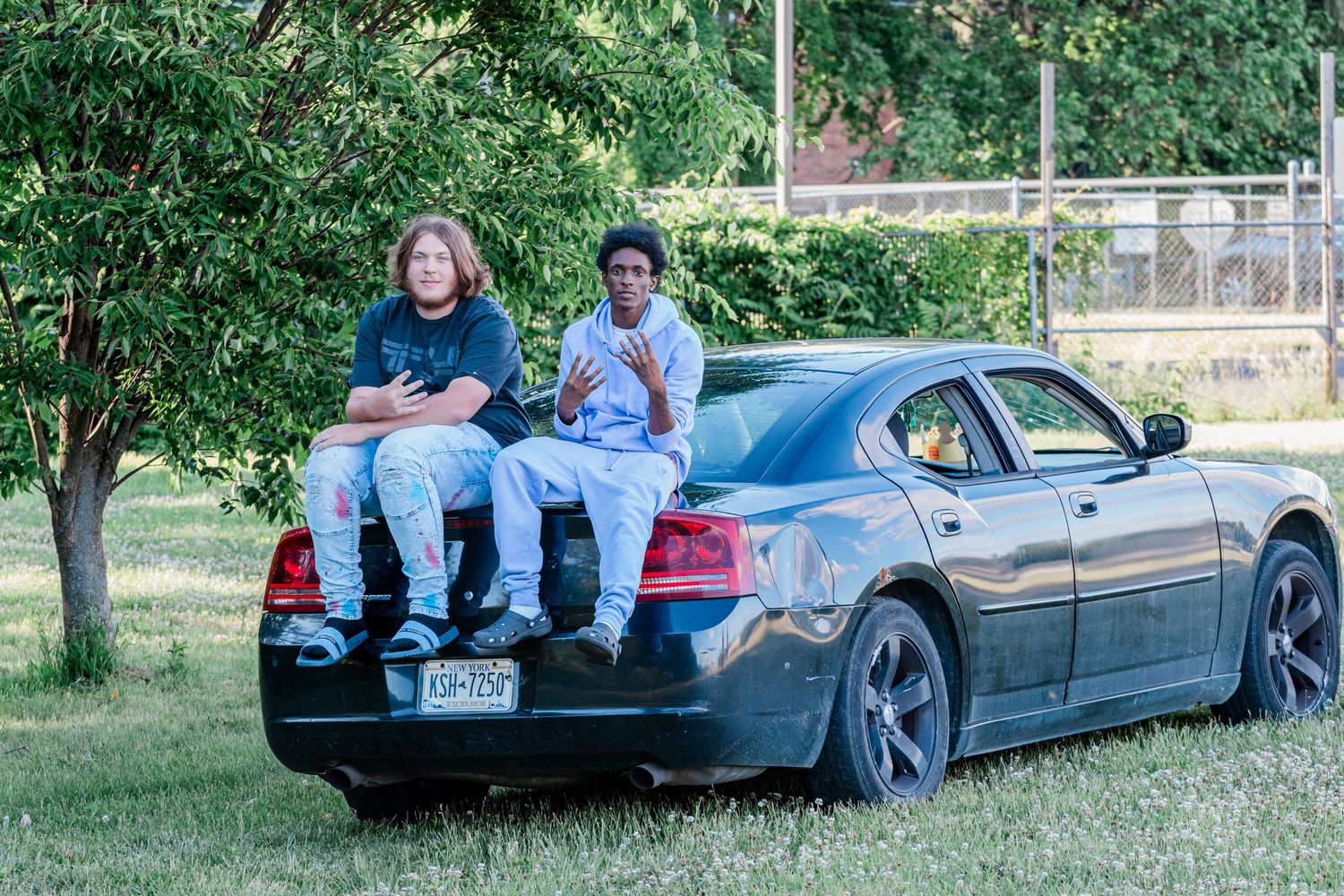 Two graduating seniors sit on the trunk of a dark sedan parked on grass beside a tree for a casual senior portrait.