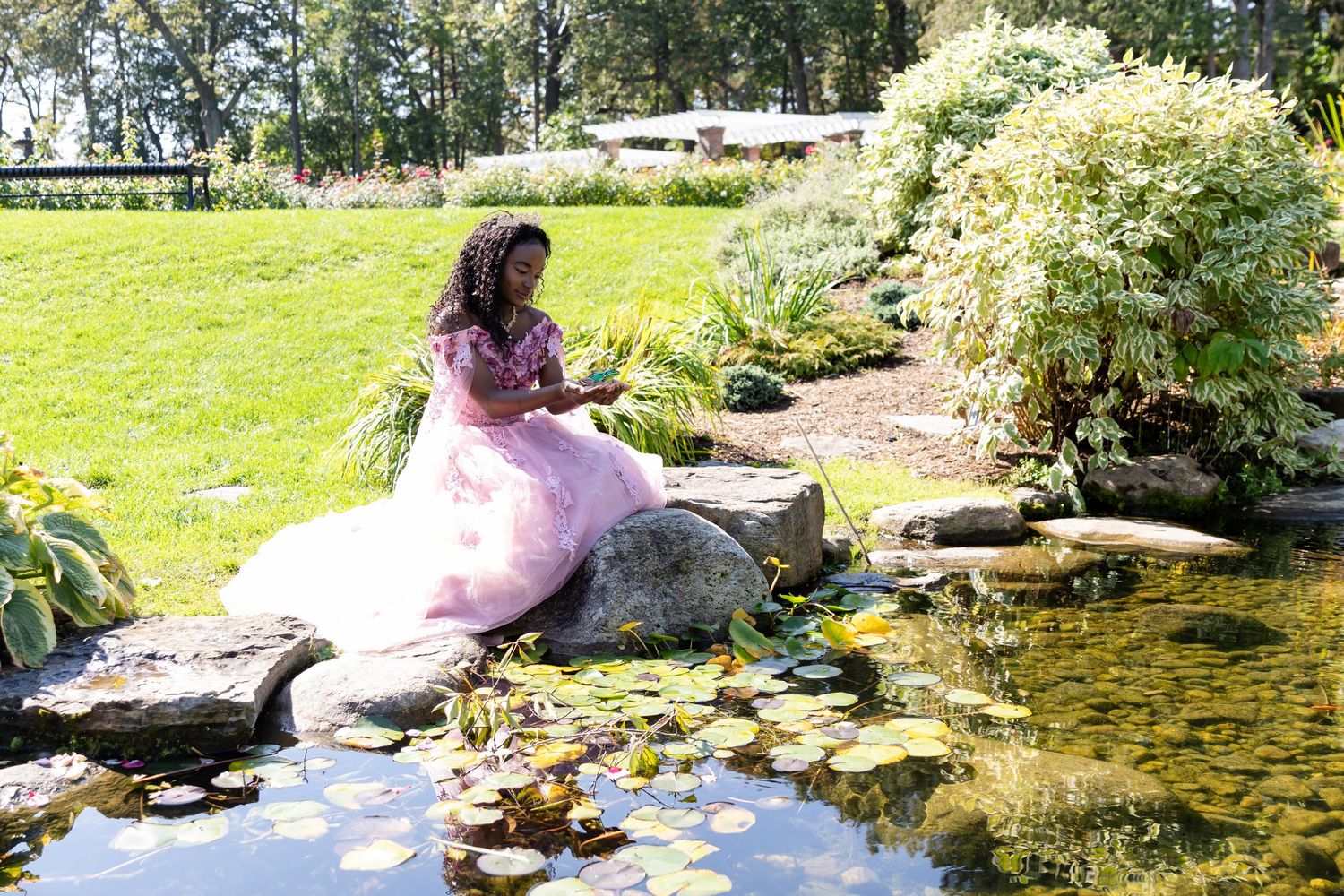 Young woman in a pink ball gown sits on a rock beside a lily pond in a landscaped garden.