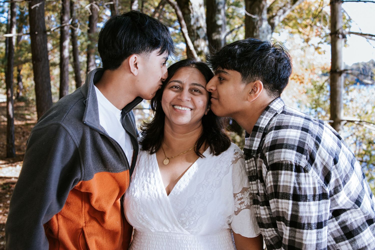 Two teens kiss a smiling woman in a white dress on both cheeks in a sunlit wooded outdoor family portrait.