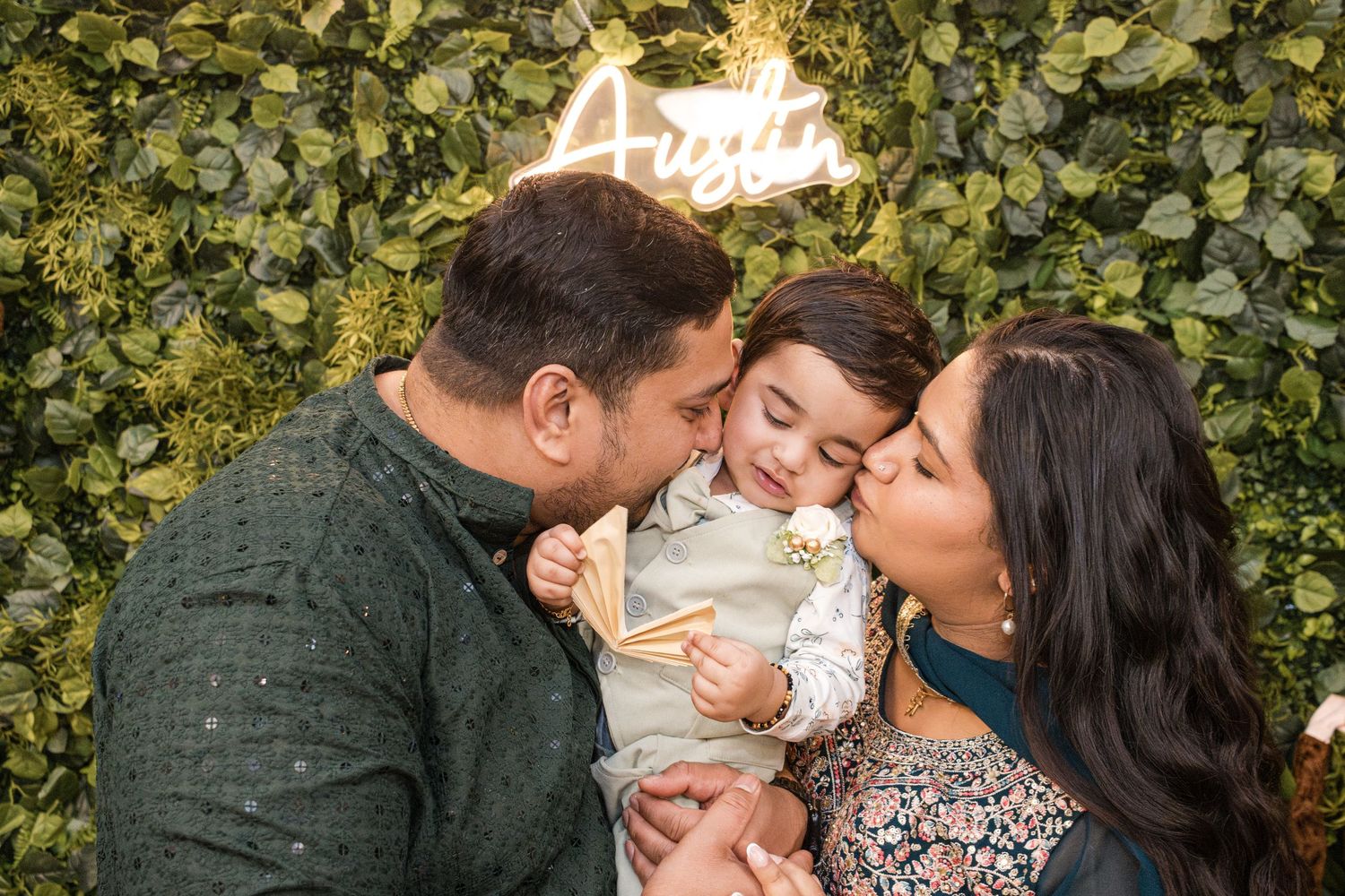 A mother and father kiss their toddler, who holds a snack, in front of a green leaf wall and neon Austin sign.