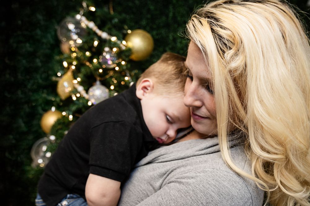 Blonde woman gently holds a sleeping toddler in a black shirt in front of a Christmas tree with gold ornaments and lights.