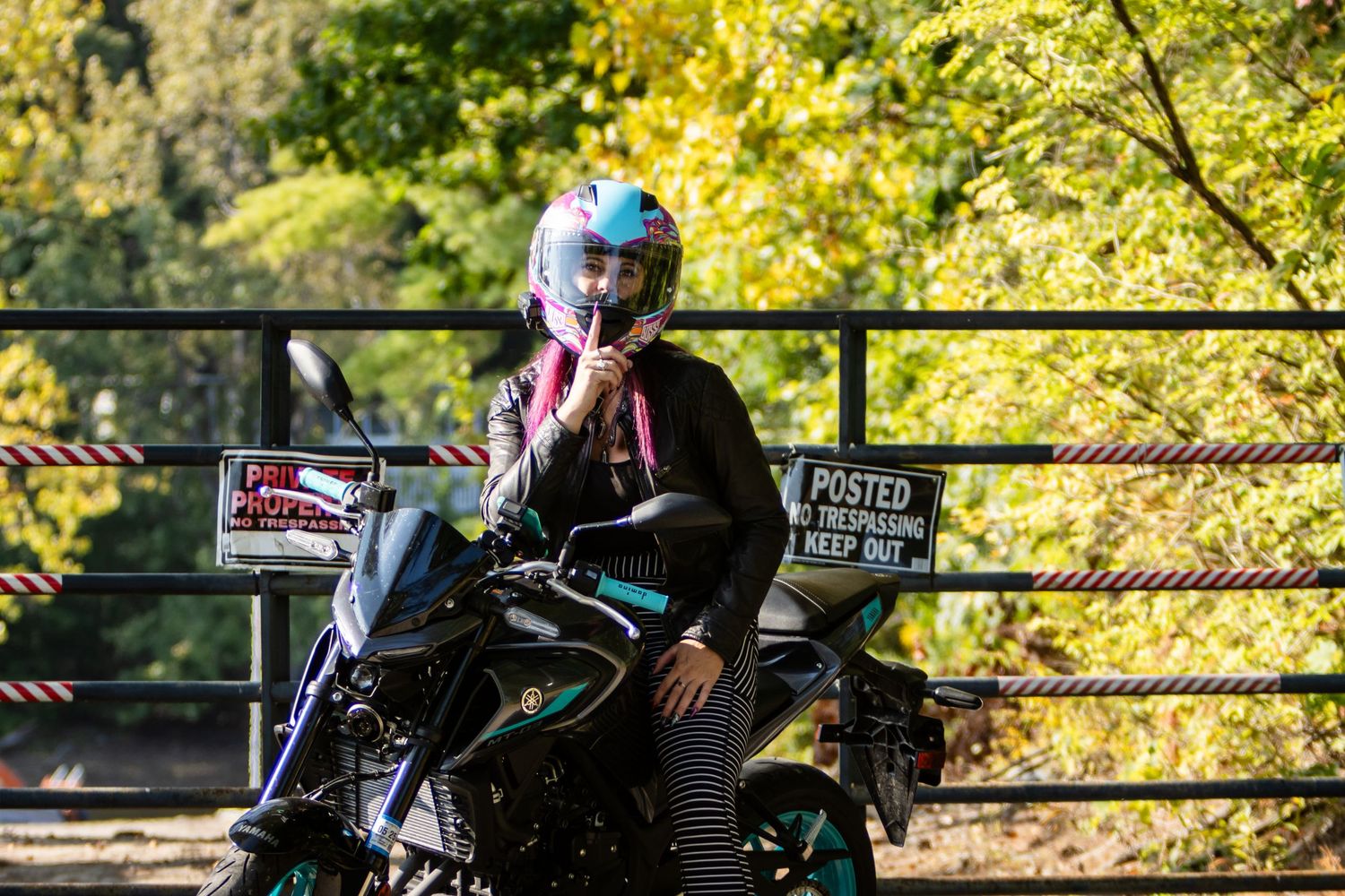 Motorcyclist in a pink and teal helmet and black jacket sits on a motorcycle beside a posted no trespassing sign and fall foliage.