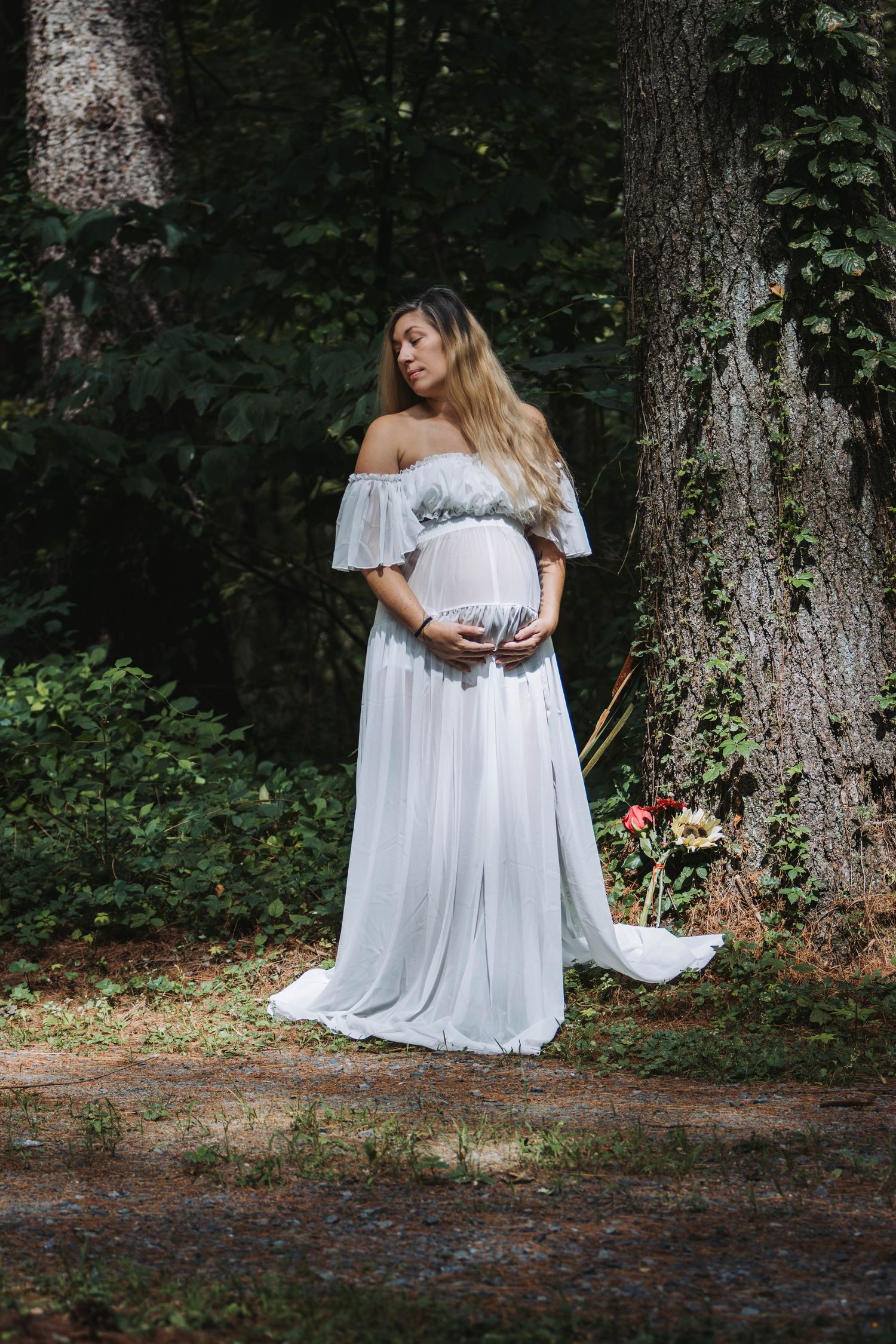 Pregnant woman in a white off-shoulder gown cradles her belly among tall trees in a forest.