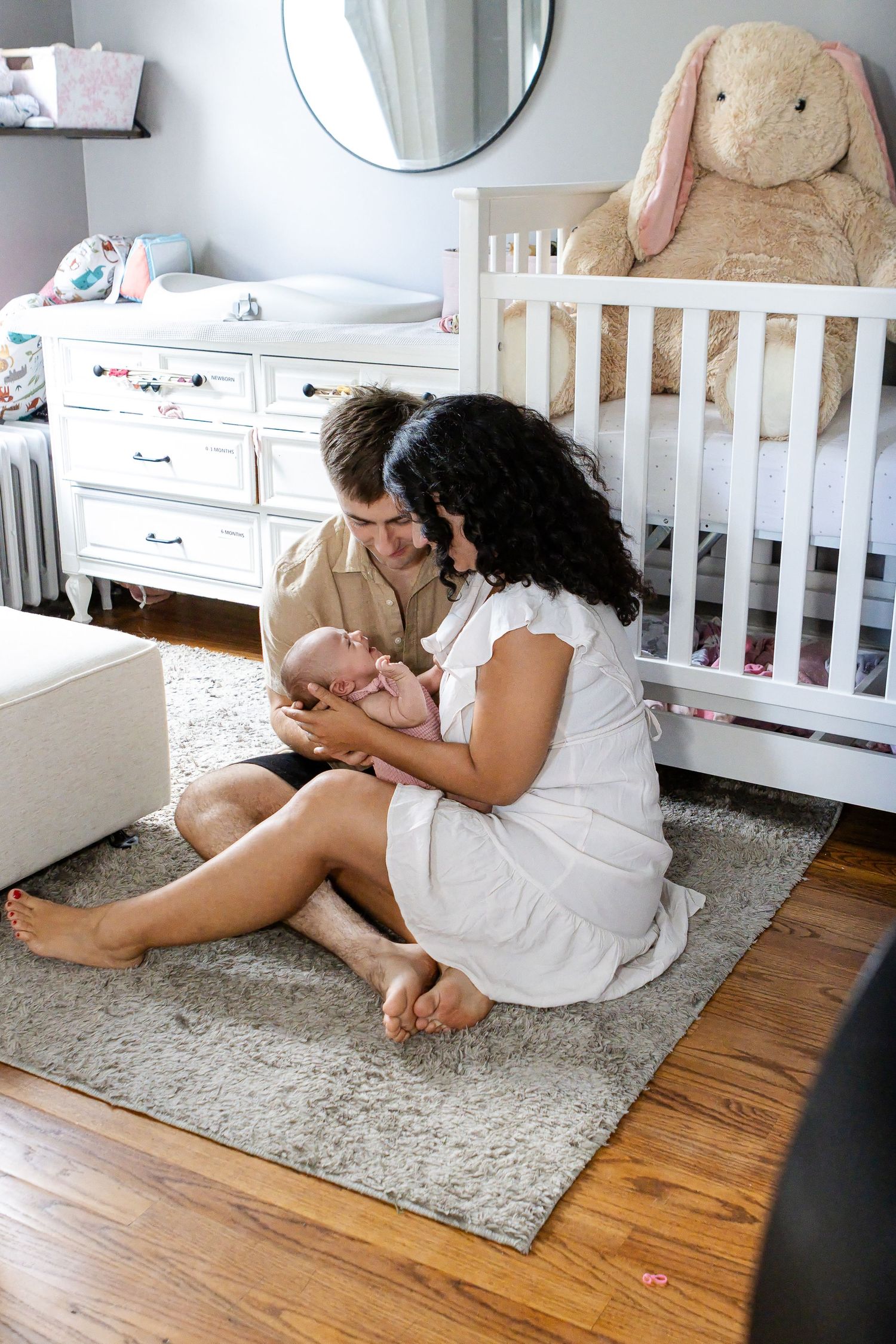 New parents sit on a gray rug in a cozy nursery, cradling their newborn baby near a white crib with a plush bunny.