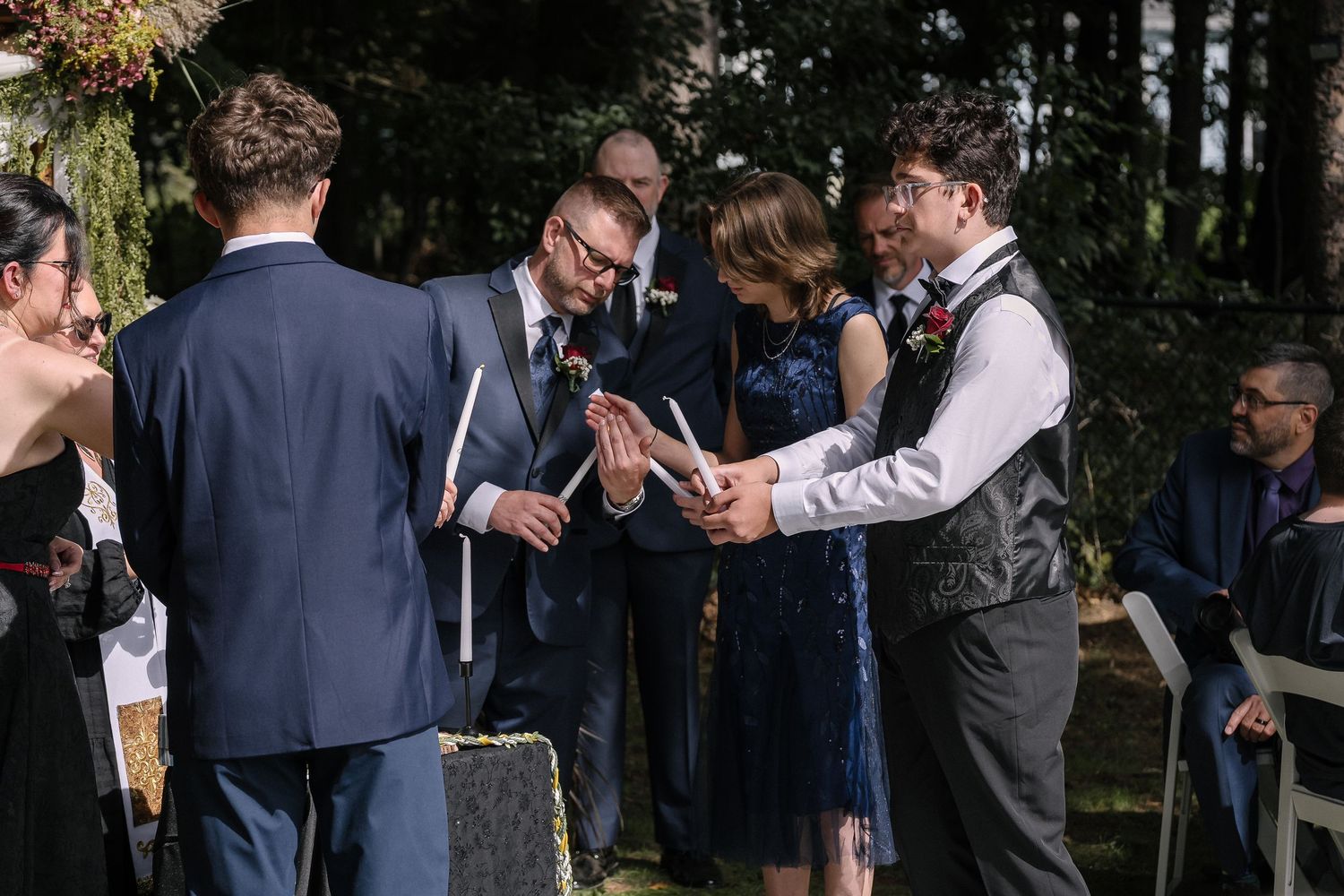 Wedding guests and the couple stand together during an outdoor ceremony, with a man in a vest and glasses reading beside the bride.