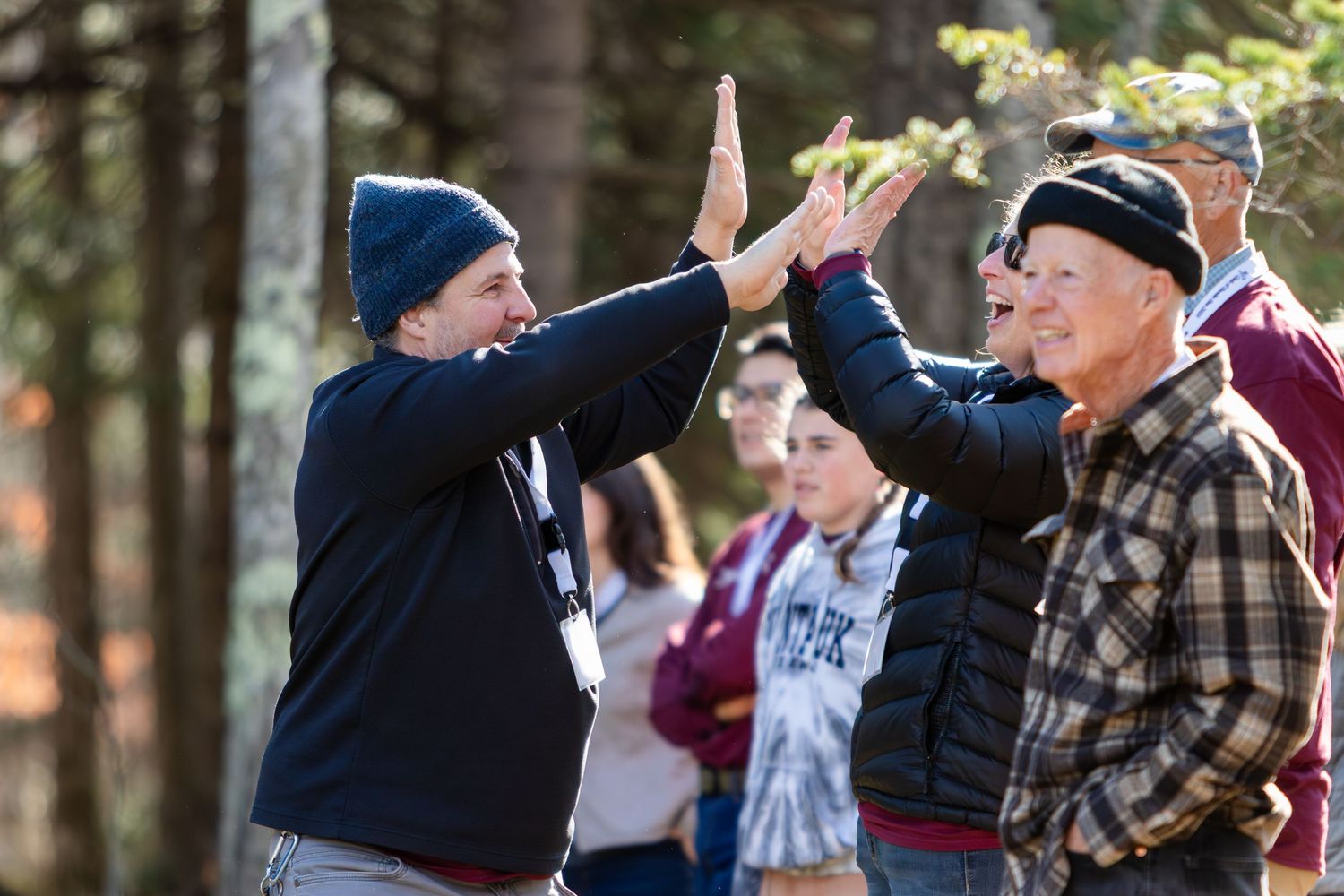 Two men in knit caps raise their hands in a high-five while a group of onlookers watches in a sunlit wooded outdoor setting.
