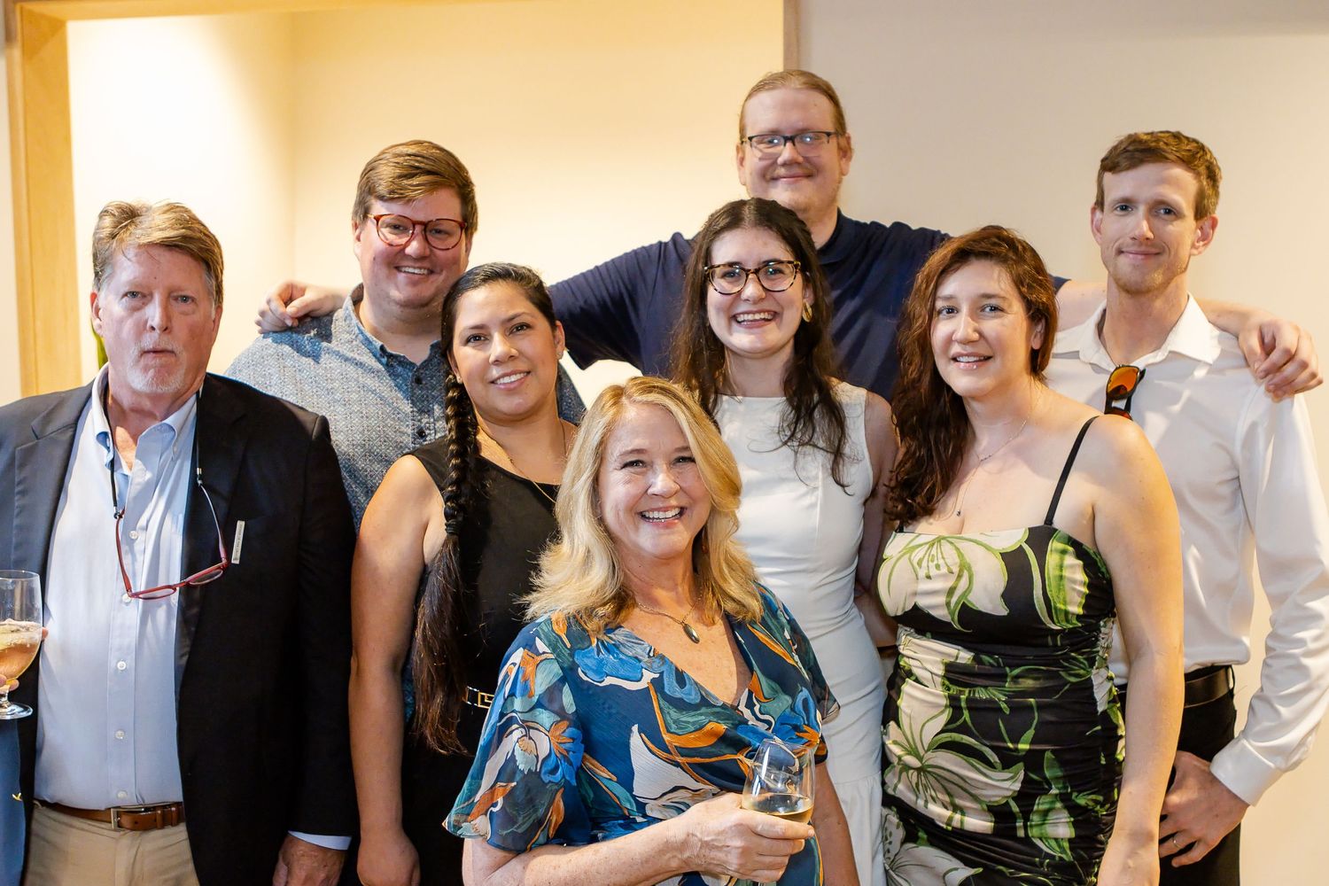 Group of eight smiling friends and colleagues in casual and formal attire posing closely together at an indoor social gathering with drinks.