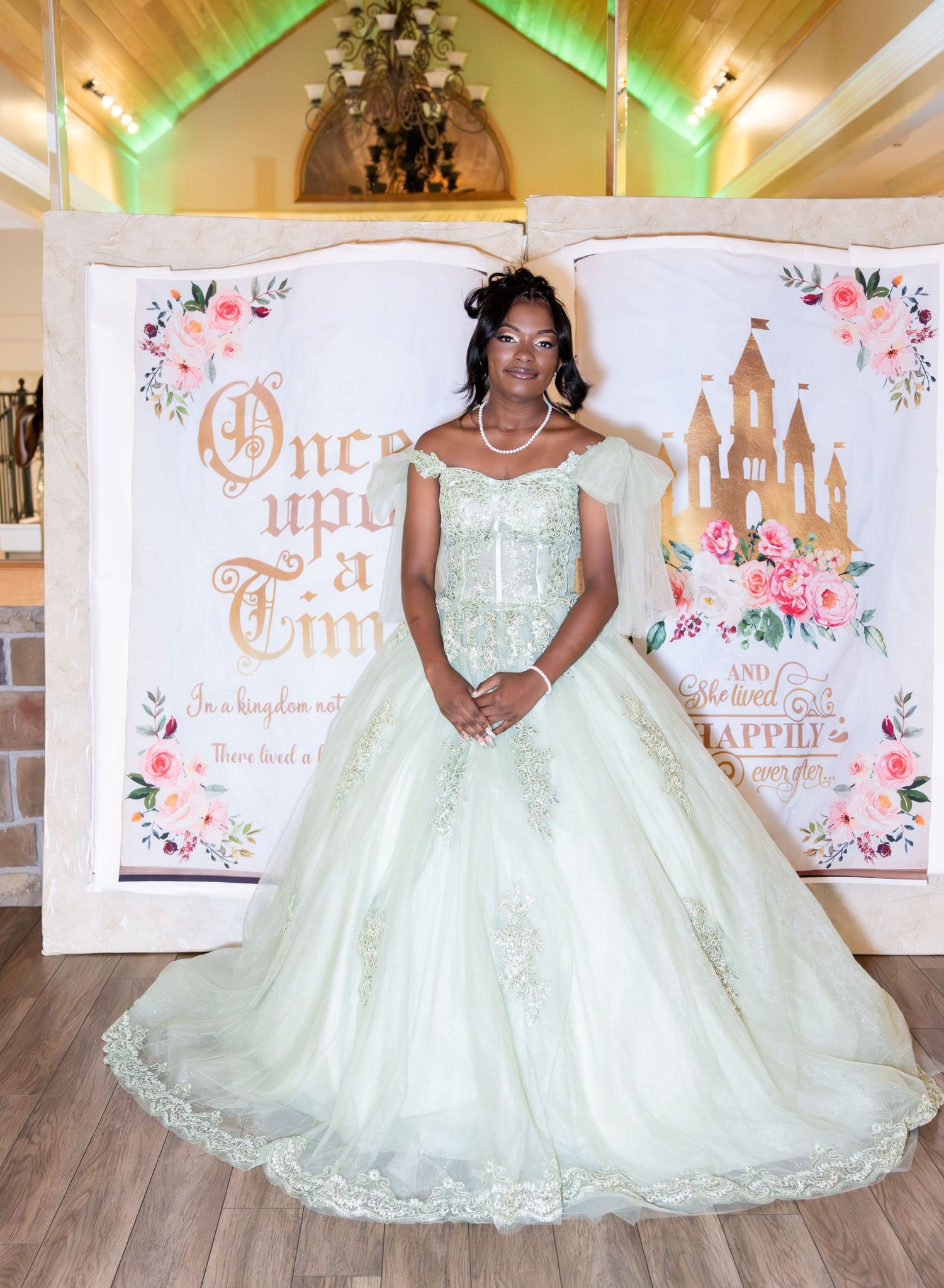 Young woman in a sage green quinceañera ball gown poses before a fairytale backdrop reading Once upon a Time with a castle and pink roses.