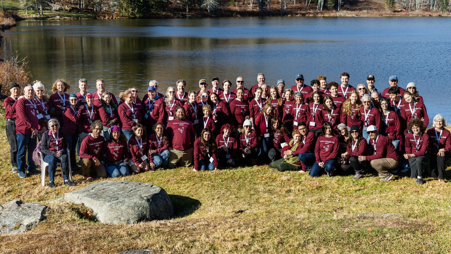Large group of people in matching maroon jackets posing together on a grassy lakeside bank with trees and calm water behind them.
