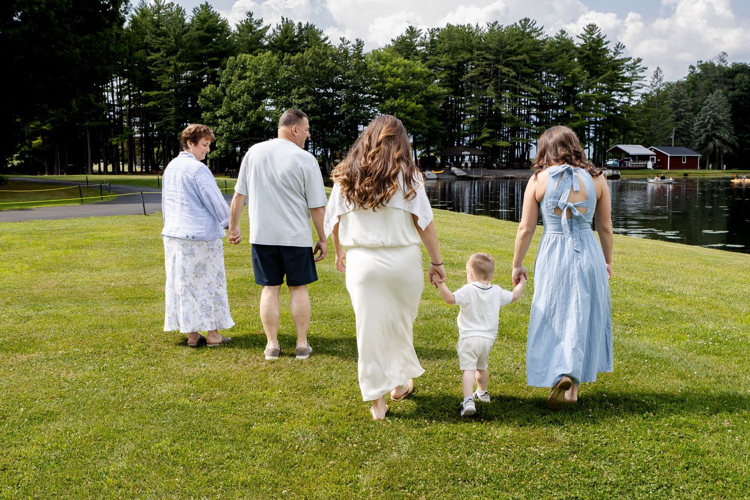 Family of five, including a toddler, walks hand in hand across a green lawn toward a lake lined with tall evergreen trees.