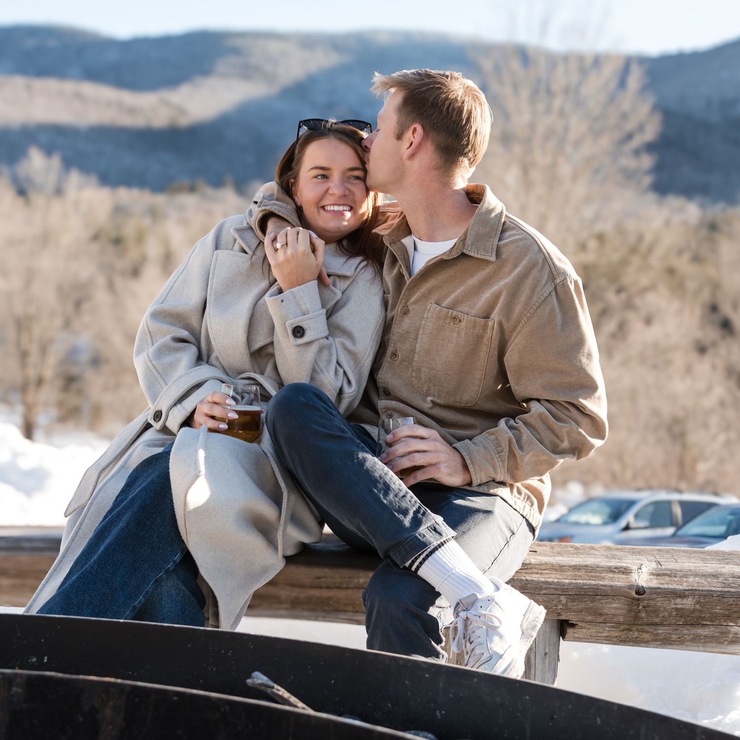 Smiling woman in a gray coat is kissed on the cheek by a man in a tan jacket as they sit on a wooden rail near snowy mountains.