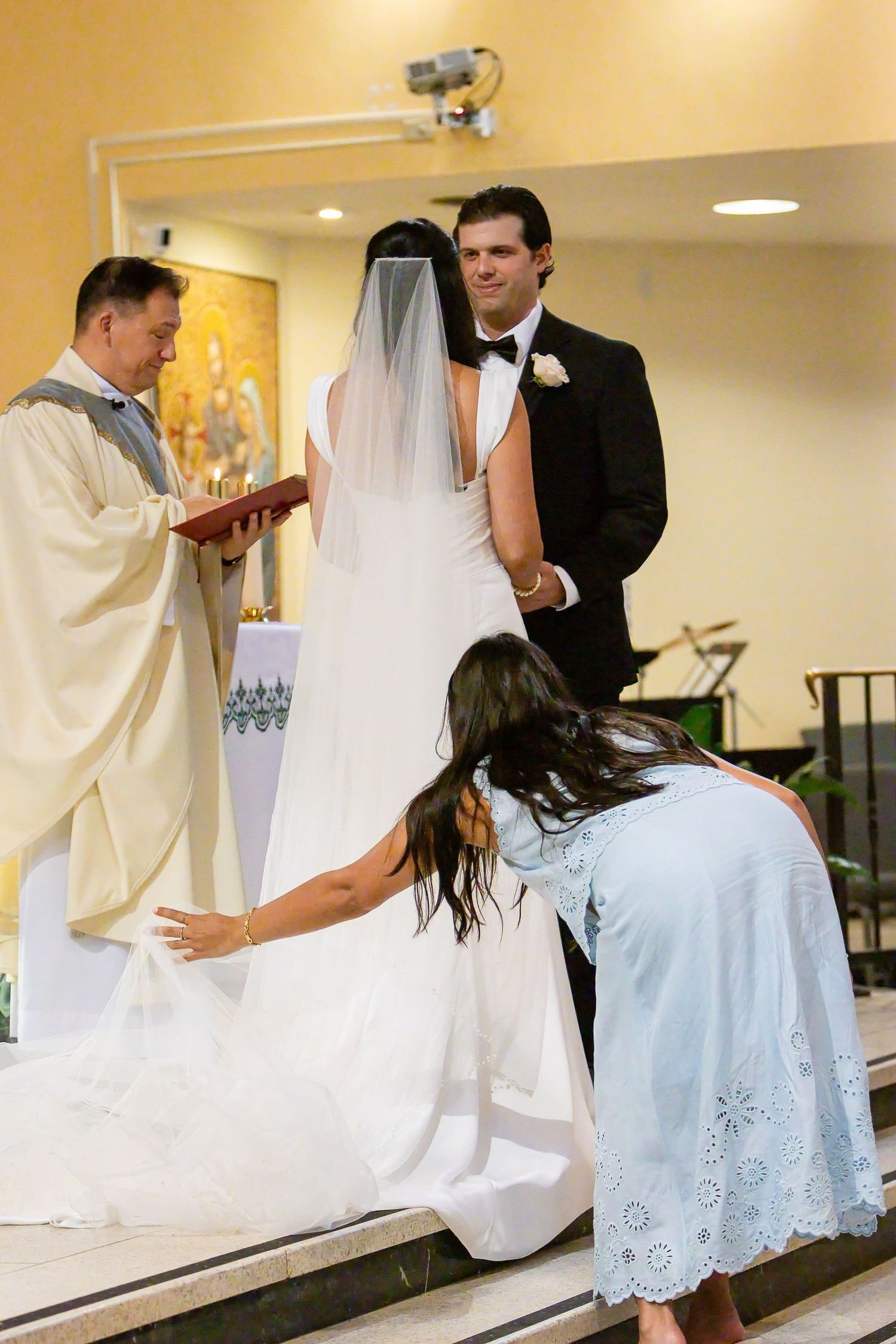 A bridesmaid bends to adjust the long veil and train of the bride as she stands before the priest and groom in a church.