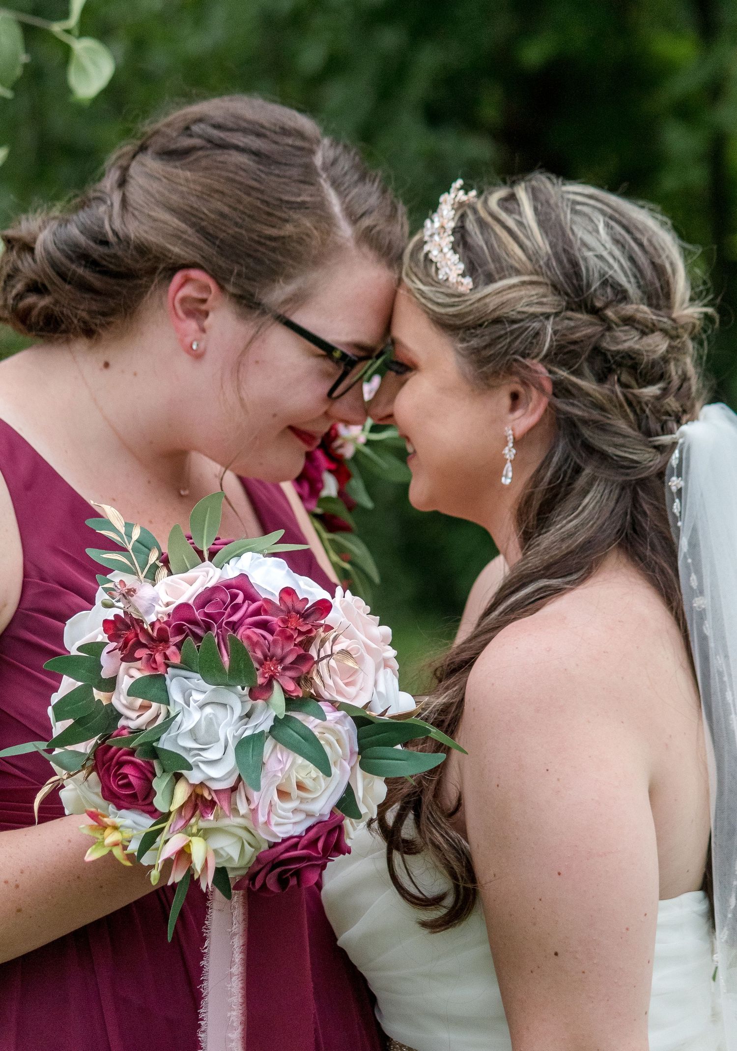 A bride and a bridesmaid in burgundy touch foreheads and smile, holding a pink and burgundy floral bouquet outdoors.