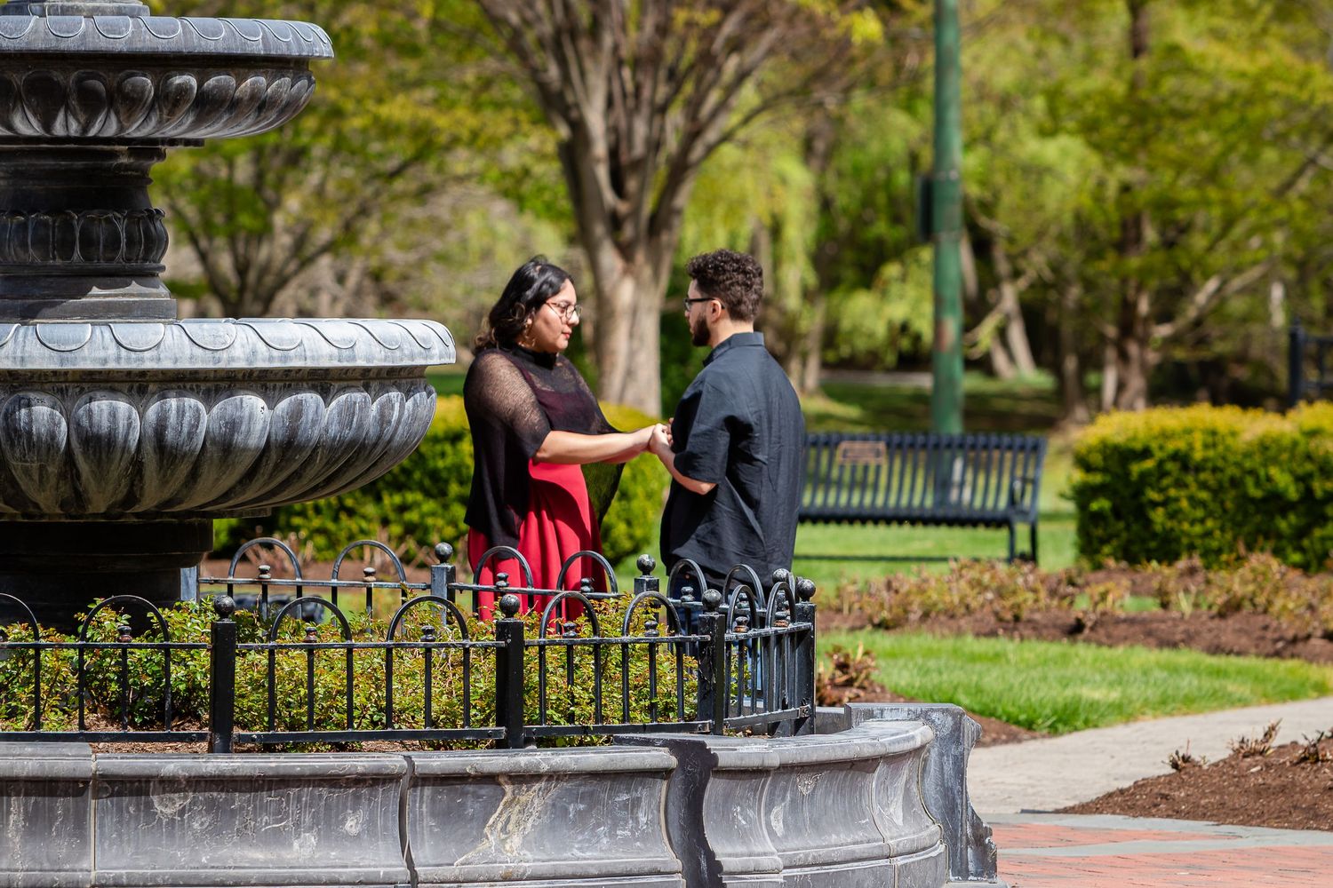 A couple stands holding hands beside a stone fountain in a lush garden park, sharing a quiet moment during a proposal.