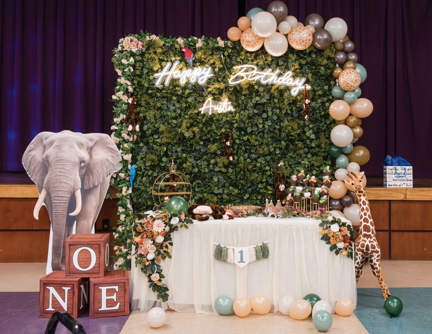 Safari baby shower backdrop with a green leaf wall, Happy Birthday neon sign, balloon garland, elephant cutout, giraffe and OXO blocks.