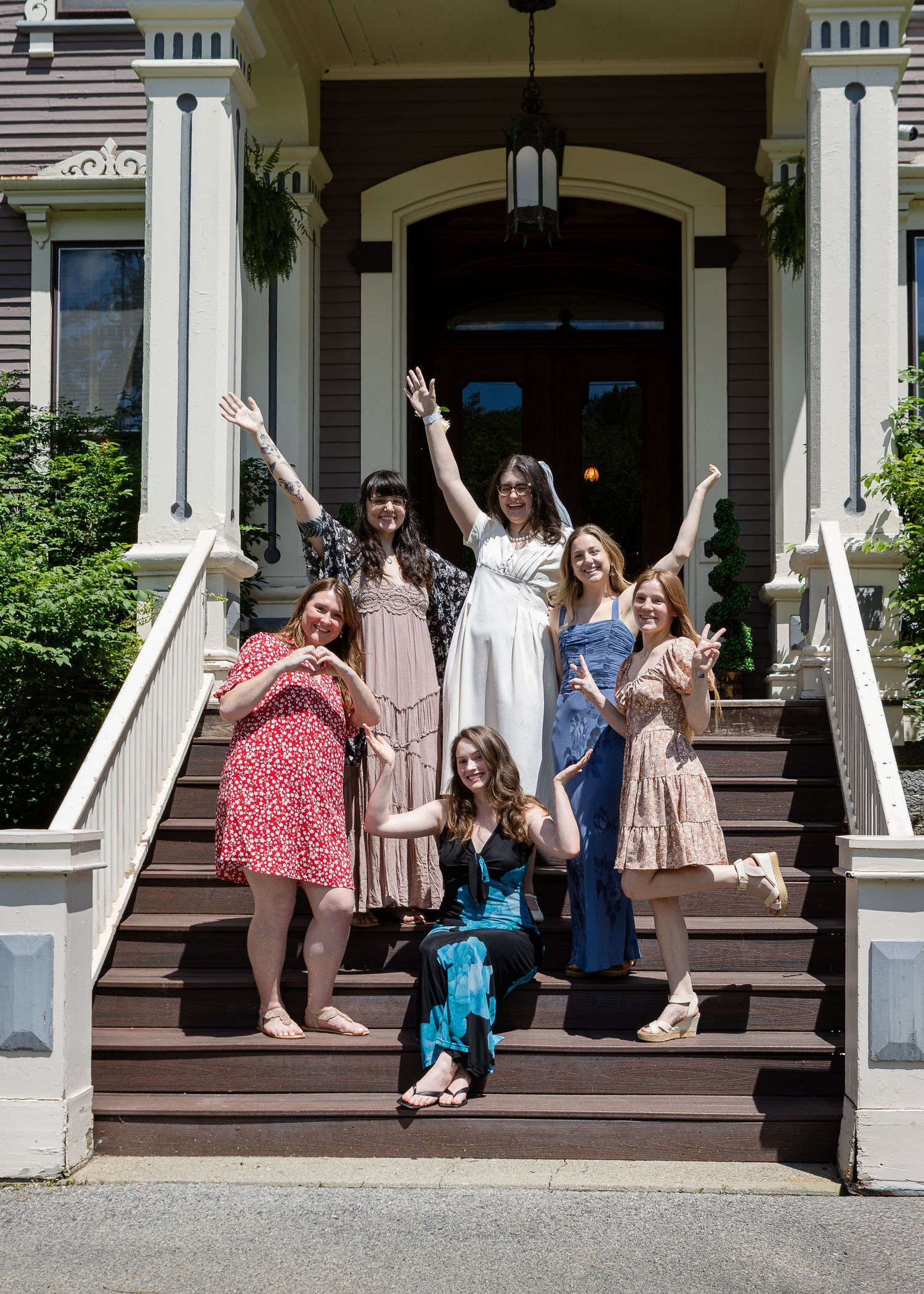 Six women in colorful summer dresses pose joyfully with raised arms on the wooden front steps of a historic house with white columns.