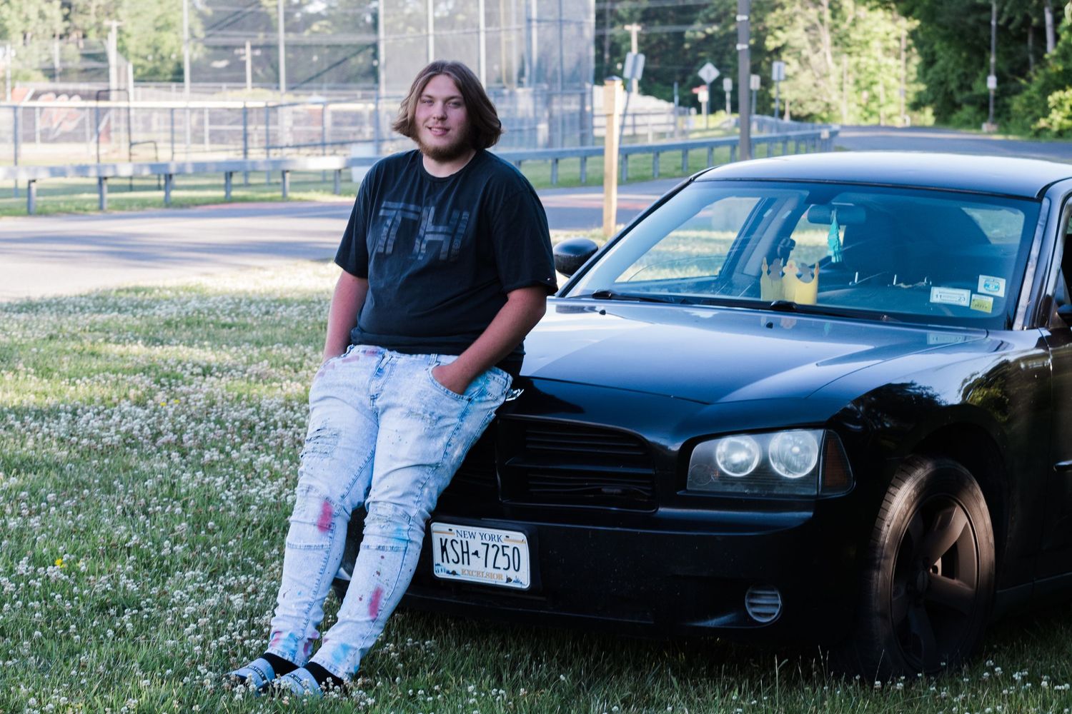 Young man in a black t-shirt and ripped jeans leans on the hood of a black sedan parked on a grassy lot.