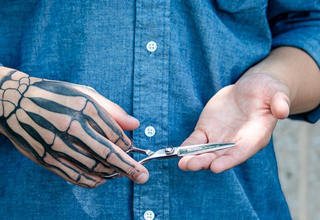 Person in a blue denim shirt holds hair-cutting shears in an open palm, with a skeleton hand tattoo on the other hand.