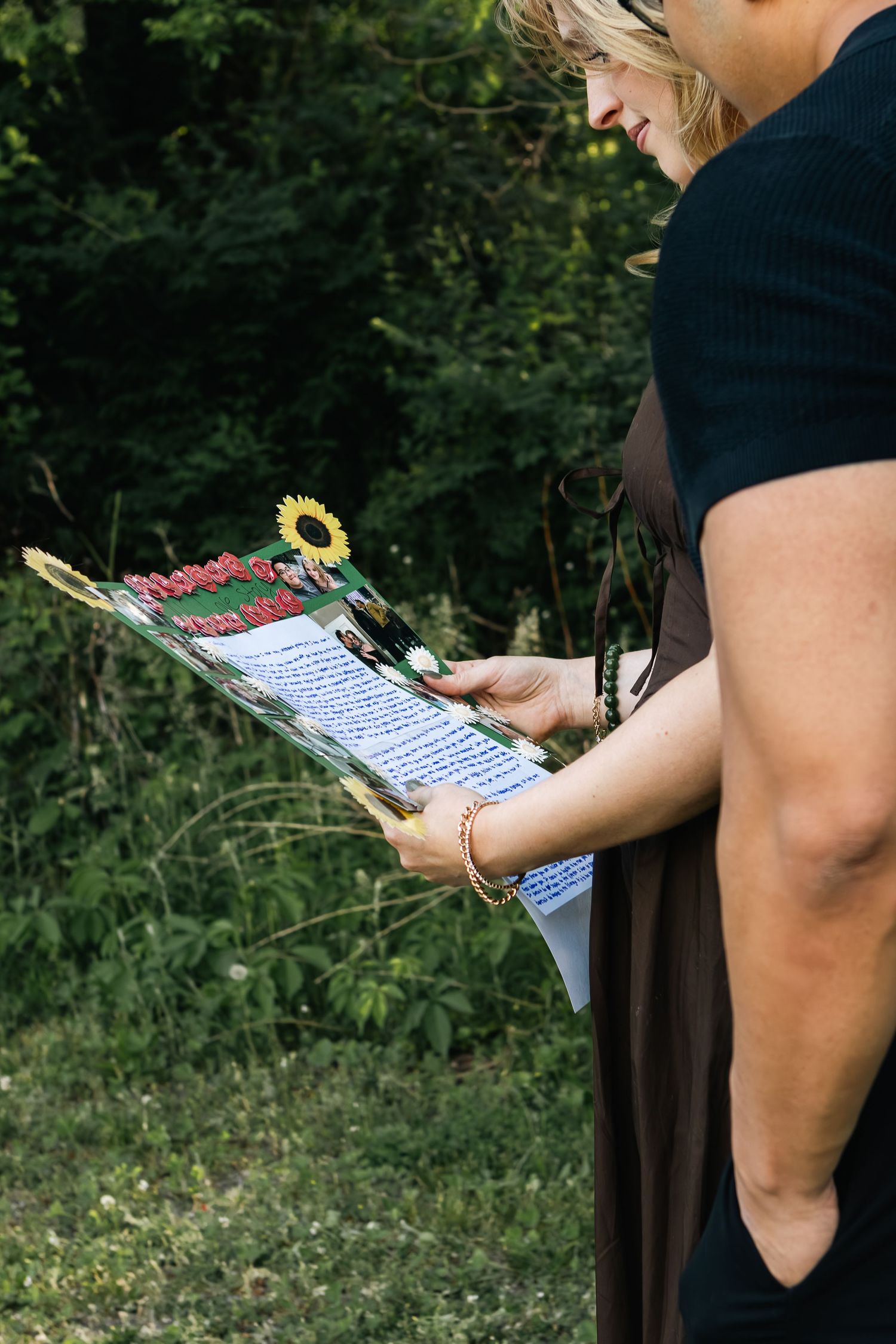 Close-up of a couple reading a handwritten letter in a decorated folder with a sunflower while standing outdoors by greenery.