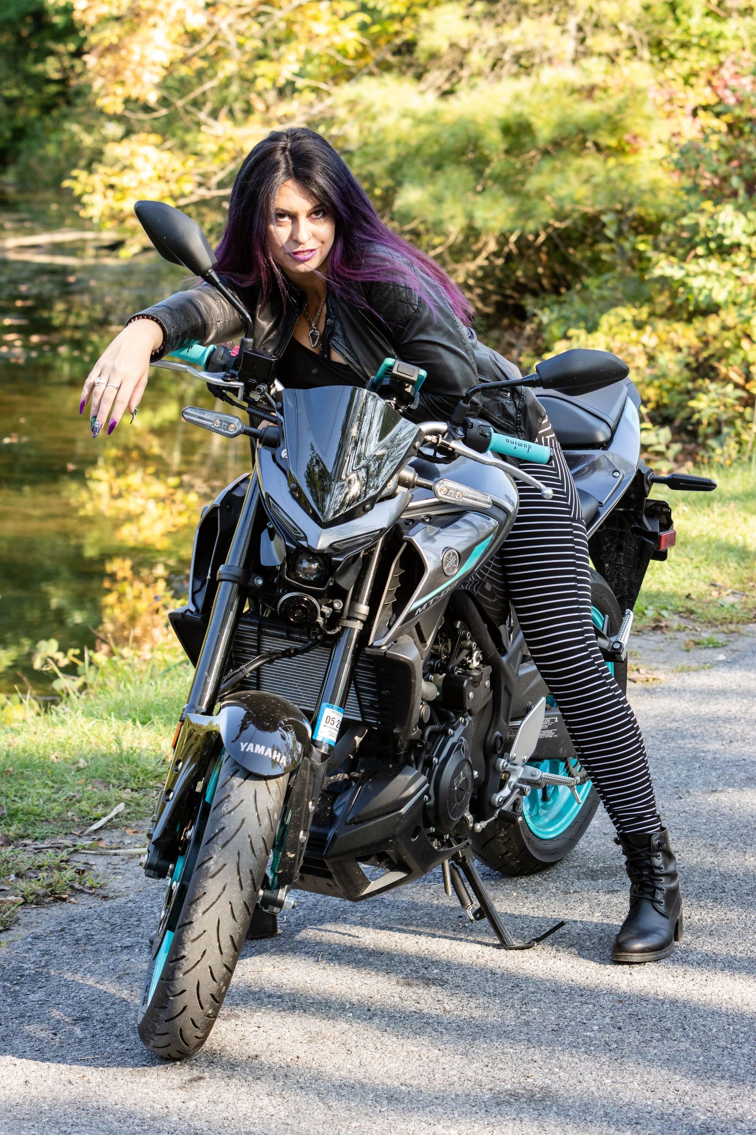 Woman in a leather jacket and striped leggings poses on a black sport motorcycle with teal wheels on a paved path near water.