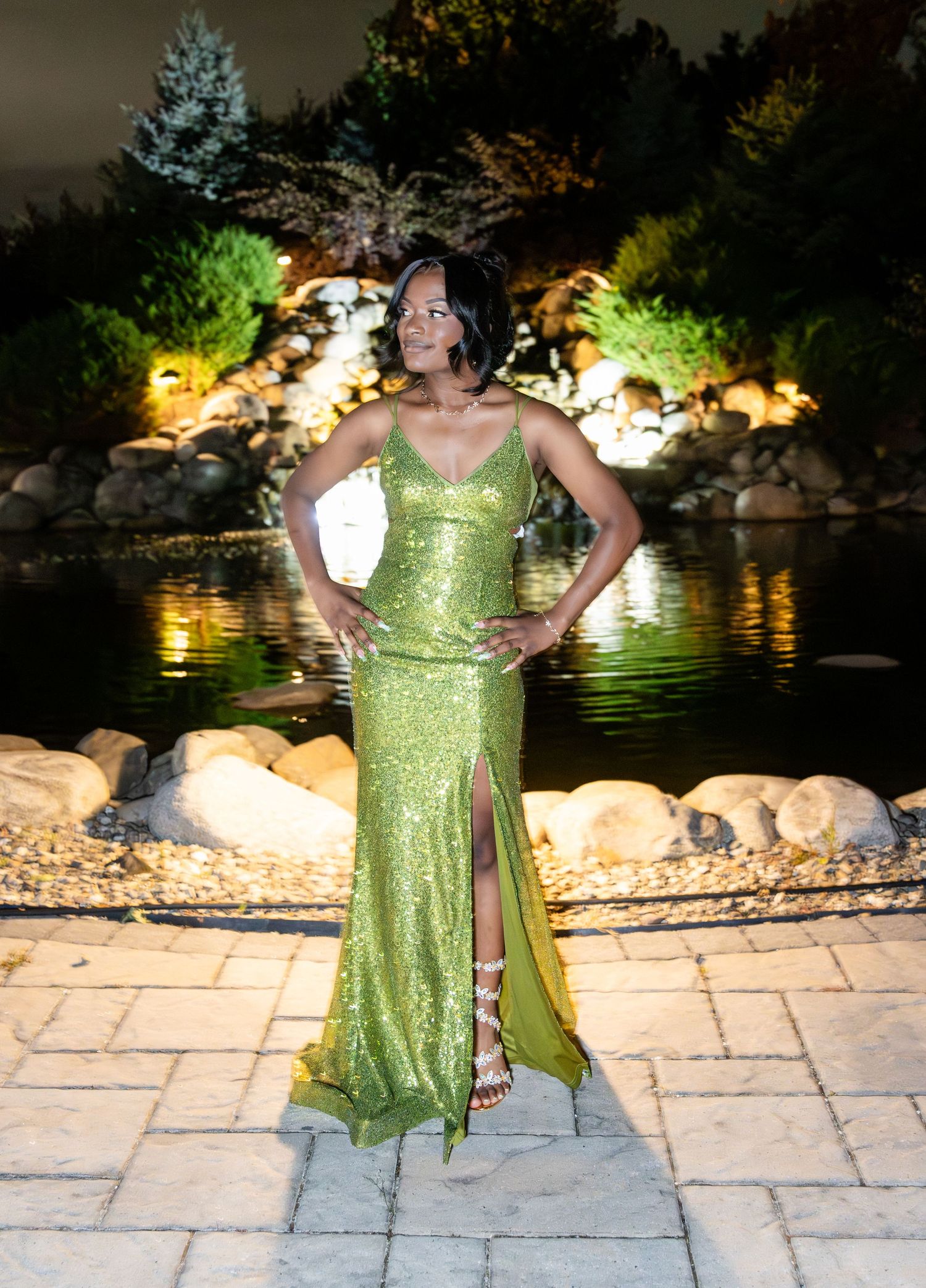 A woman in a sparkling lime green gown with a high slit poses on a stone patio beside a lit garden pond at night.