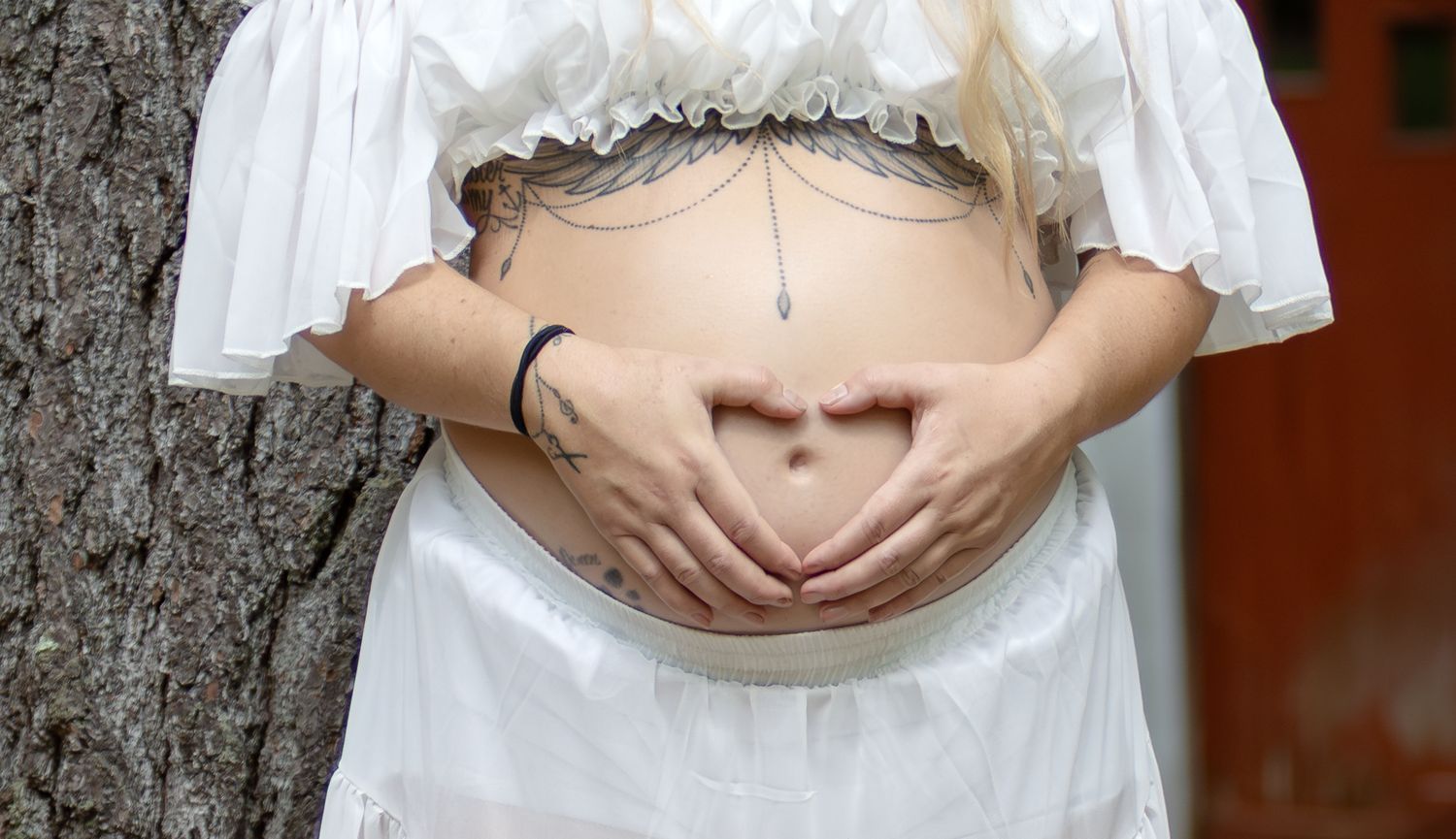 Close-up of a pregnant woman in white forming a heart shape with her hands on her baby bump.