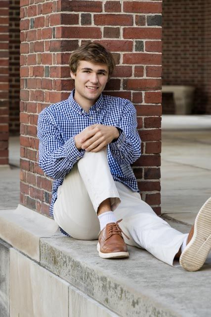 Smiling young man in a blue plaid shirt, khaki pants and brown shoes sits on a stone ledge by a brick column.