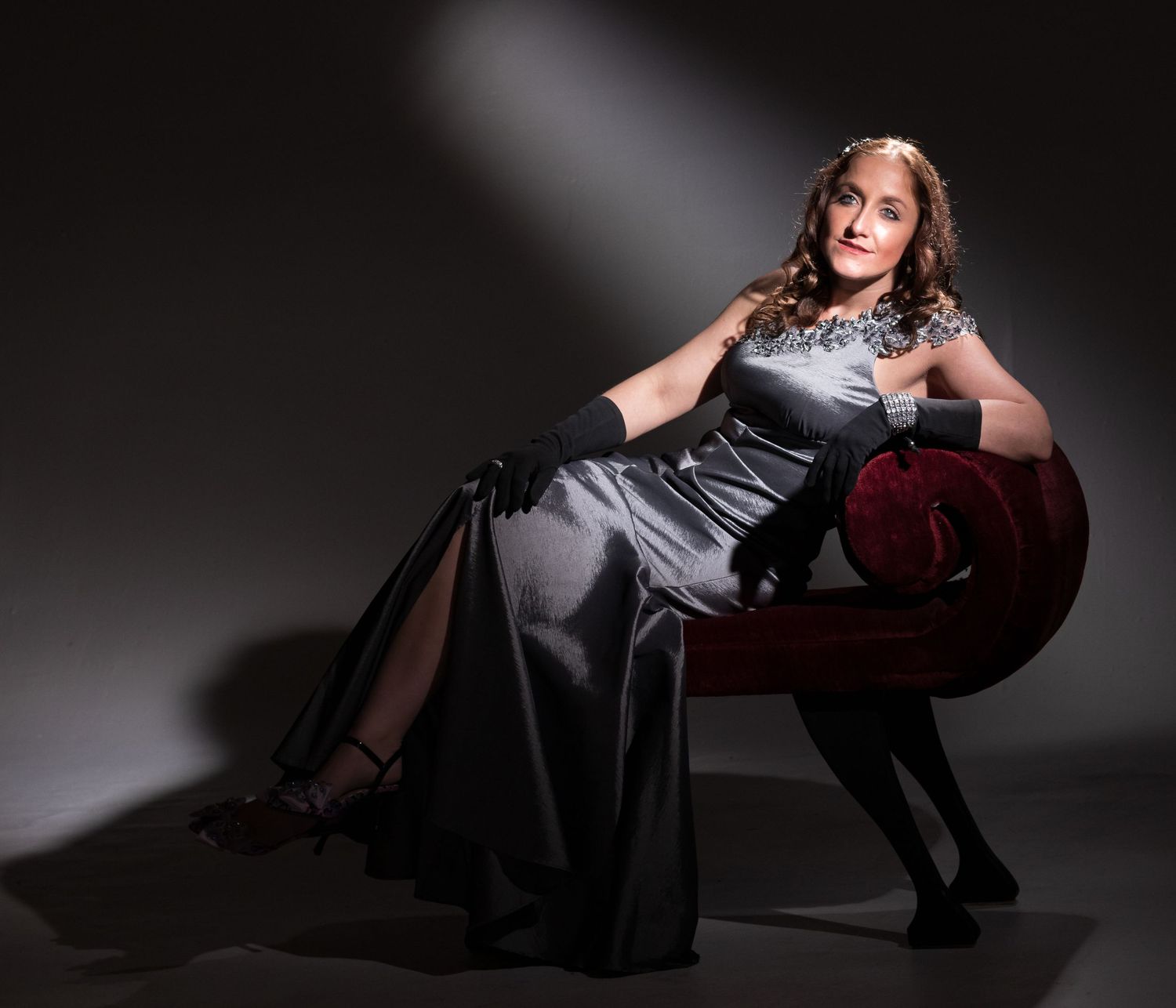 Woman in a silver satin gown and black gloves poses on a red velvet chaise lounge in a vintage-style studio portrait.