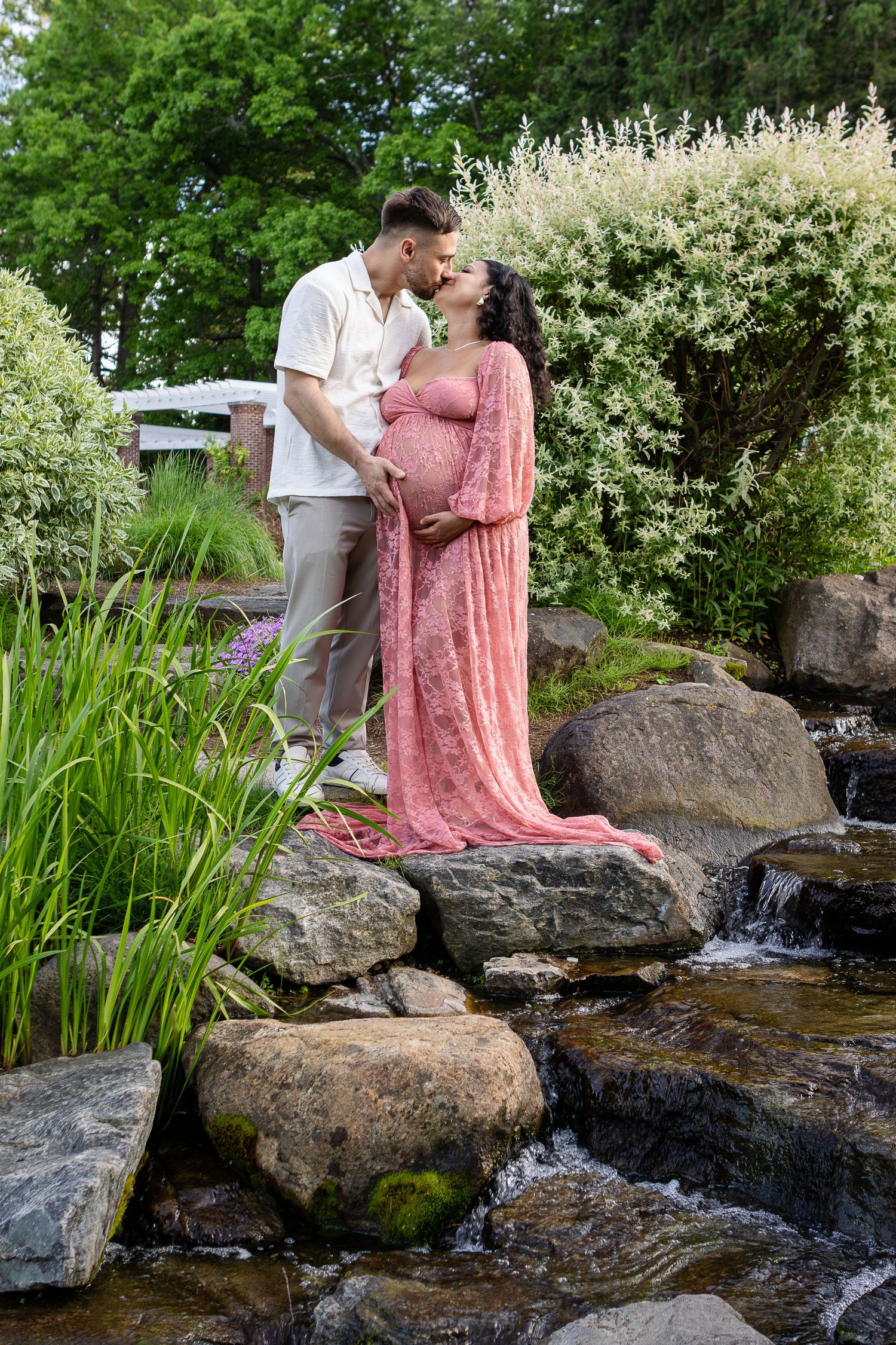 An expecting couple in a pink gown and white shirt kiss on mossy rocks beside a garden stream.