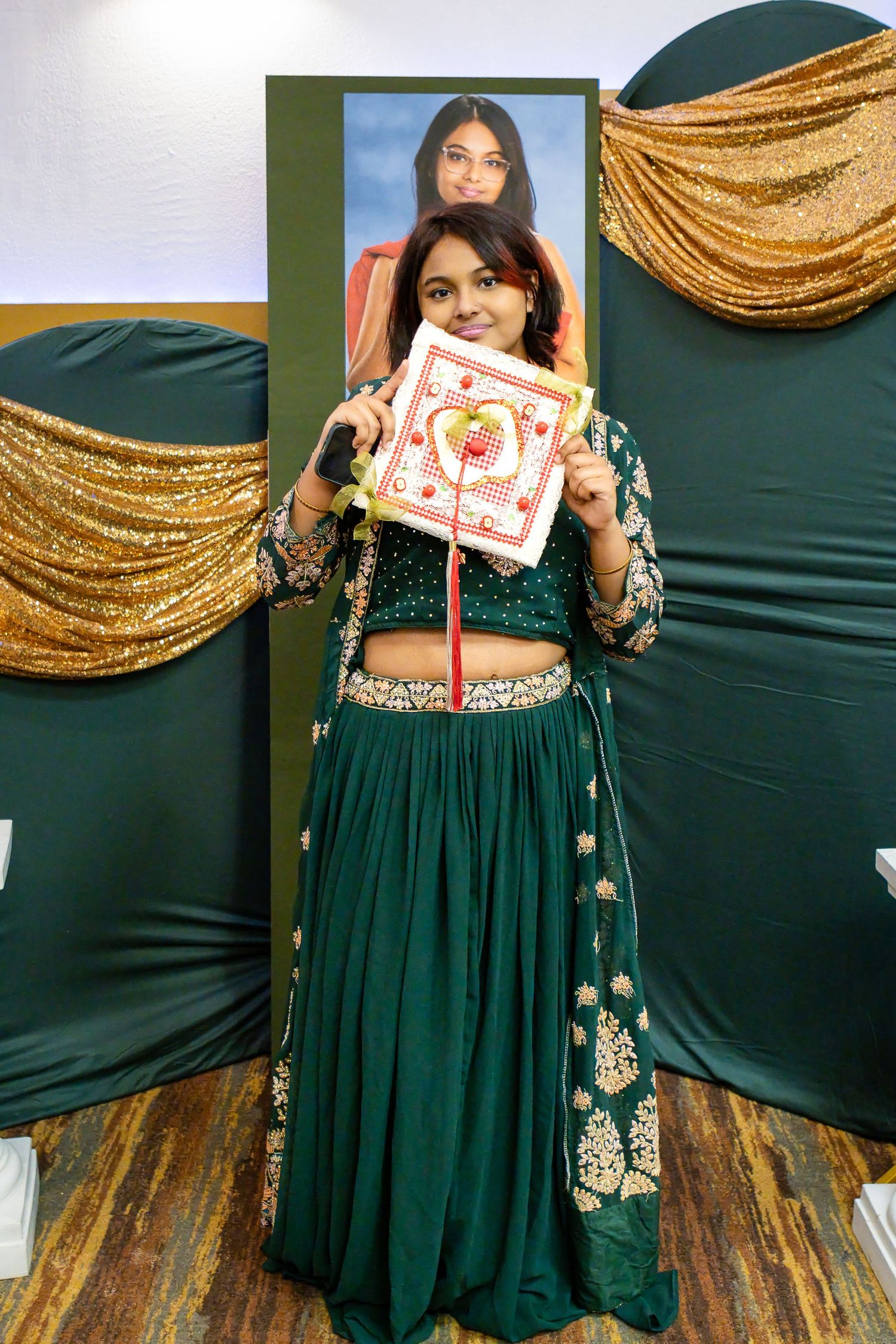 Smiling graduate in an emerald green embroidered lehenga holds a decorated graduation cap in front of a portrait and gold draped backdrop.