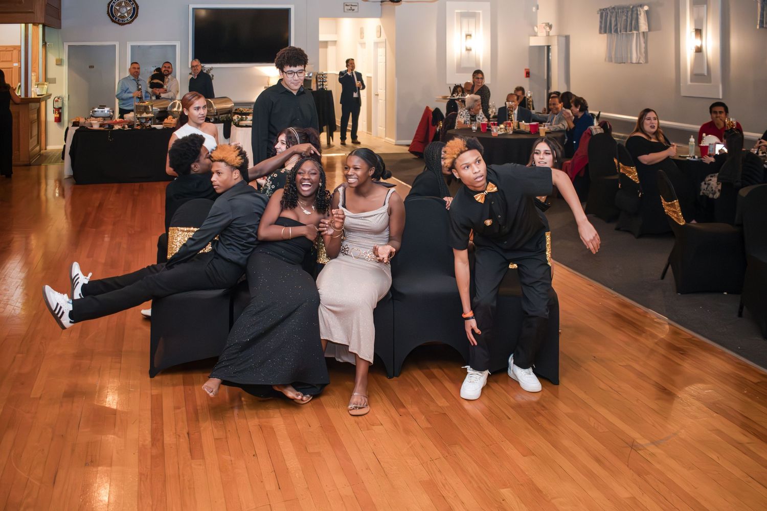 Group of people in black and white outfits gather on sofas and chairs at a social event in a banquet hall with a polished wood floor.
