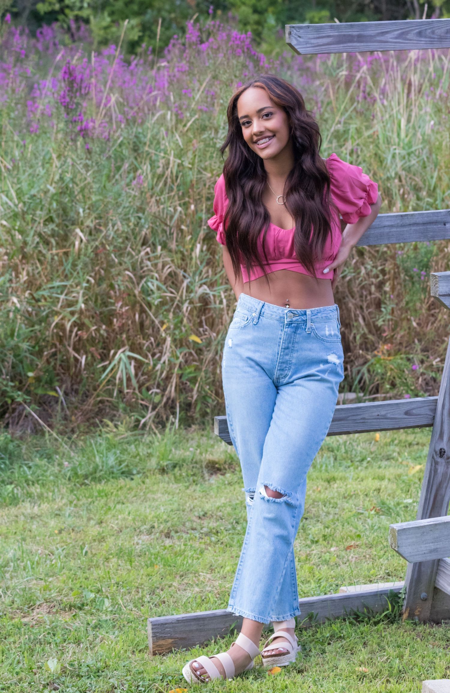 Smiling young woman in a pink top and ripped light-wash jeans leaning on a wooden fence beside tall purple wildflowers.