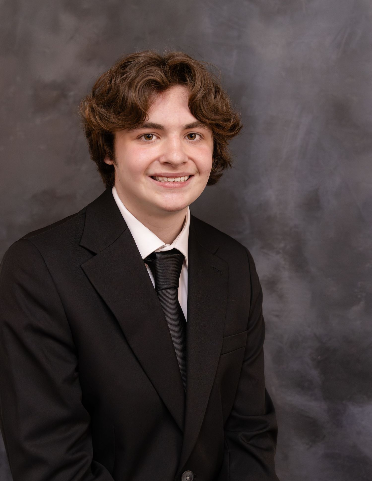 Studio portrait of a smiling teenage boy with wavy brown hair wearing a black suit and tie against a gray backdrop.