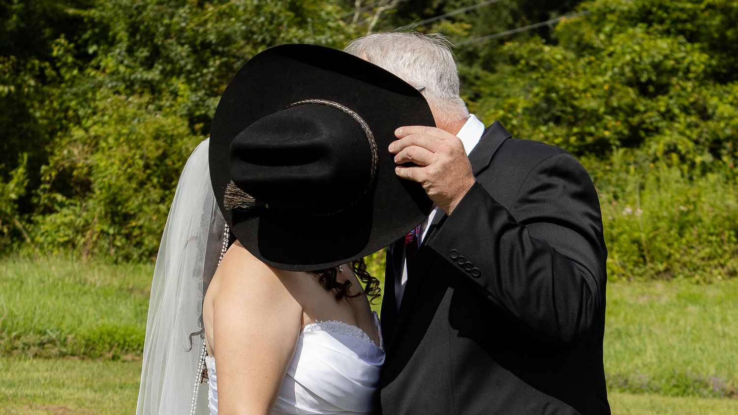 A groom in a black suit and a bride in a white gown with veil kiss behind a black cowboy hat on a sunny grassy field.