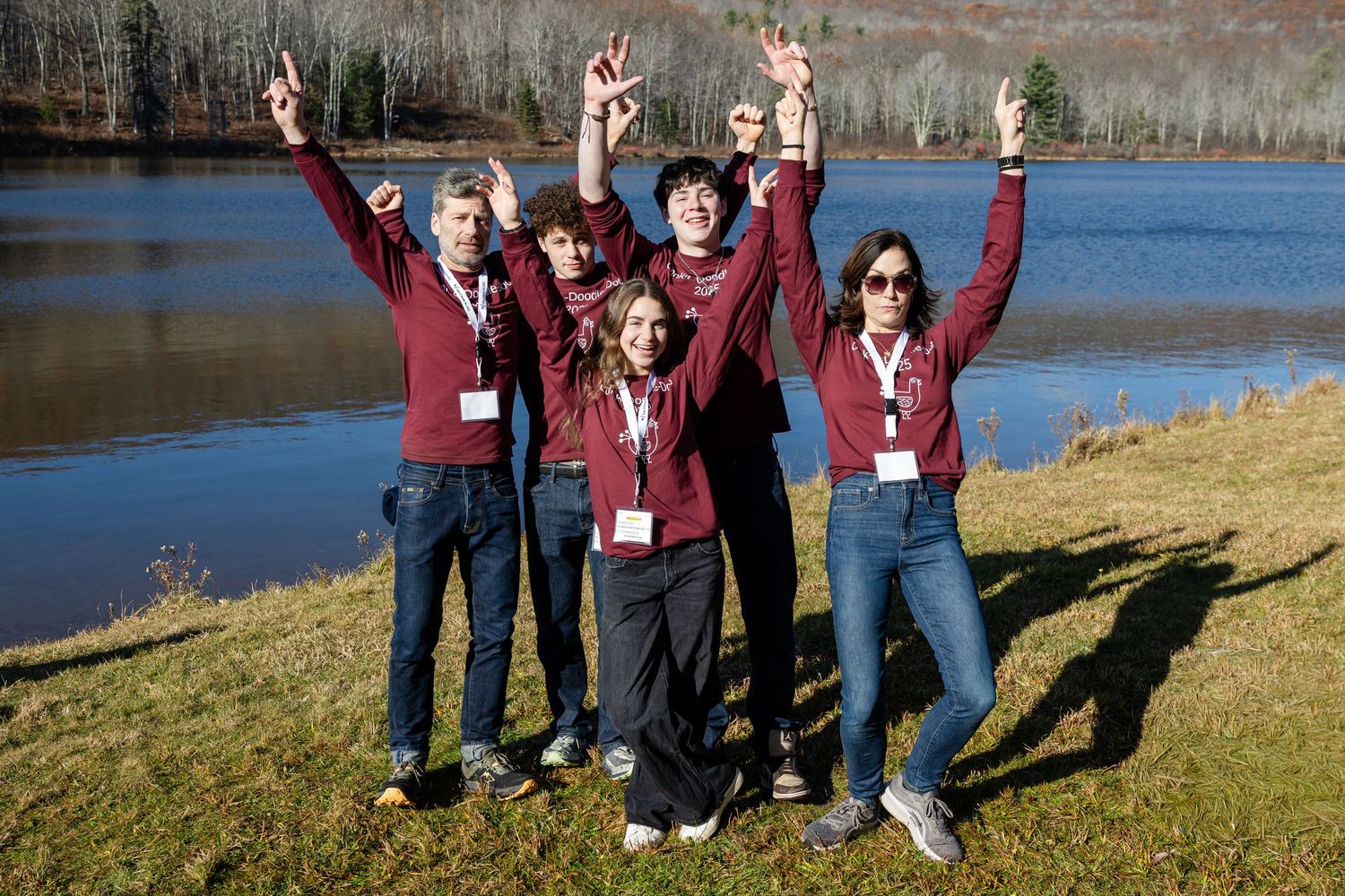 Group of smiling people in matching maroon shirts and name badges raise their arms in celebration on a grassy lakeside shore.