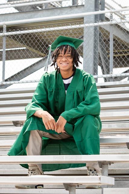 Smiling graduate in a green cap and gown sits on metal bleachers in front of a chain-link fence for a senior portrait.