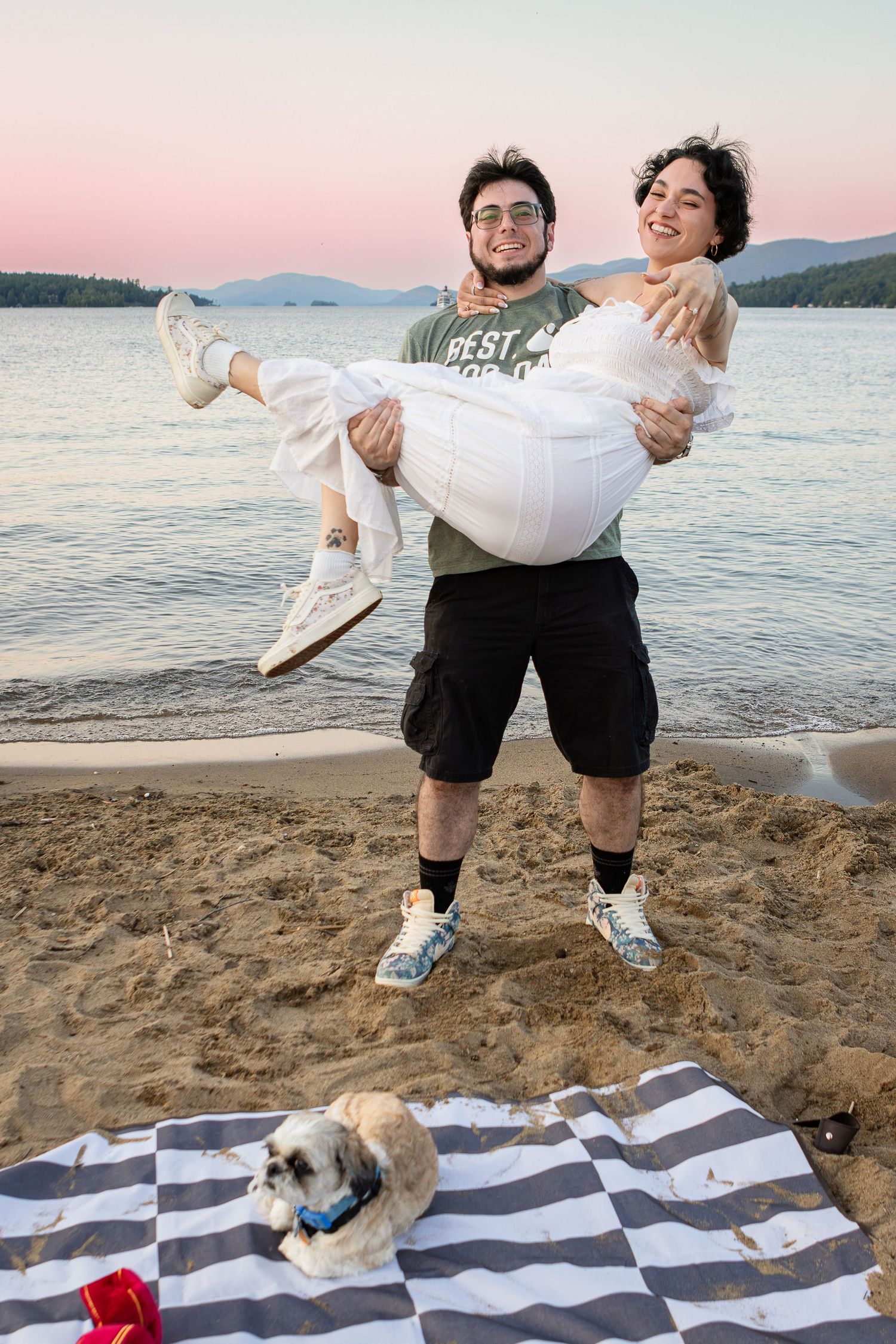 A bearded man in glasses carries a laughing woman in white on a sandy lake beach at sunset while a small dog rests on a blanket.