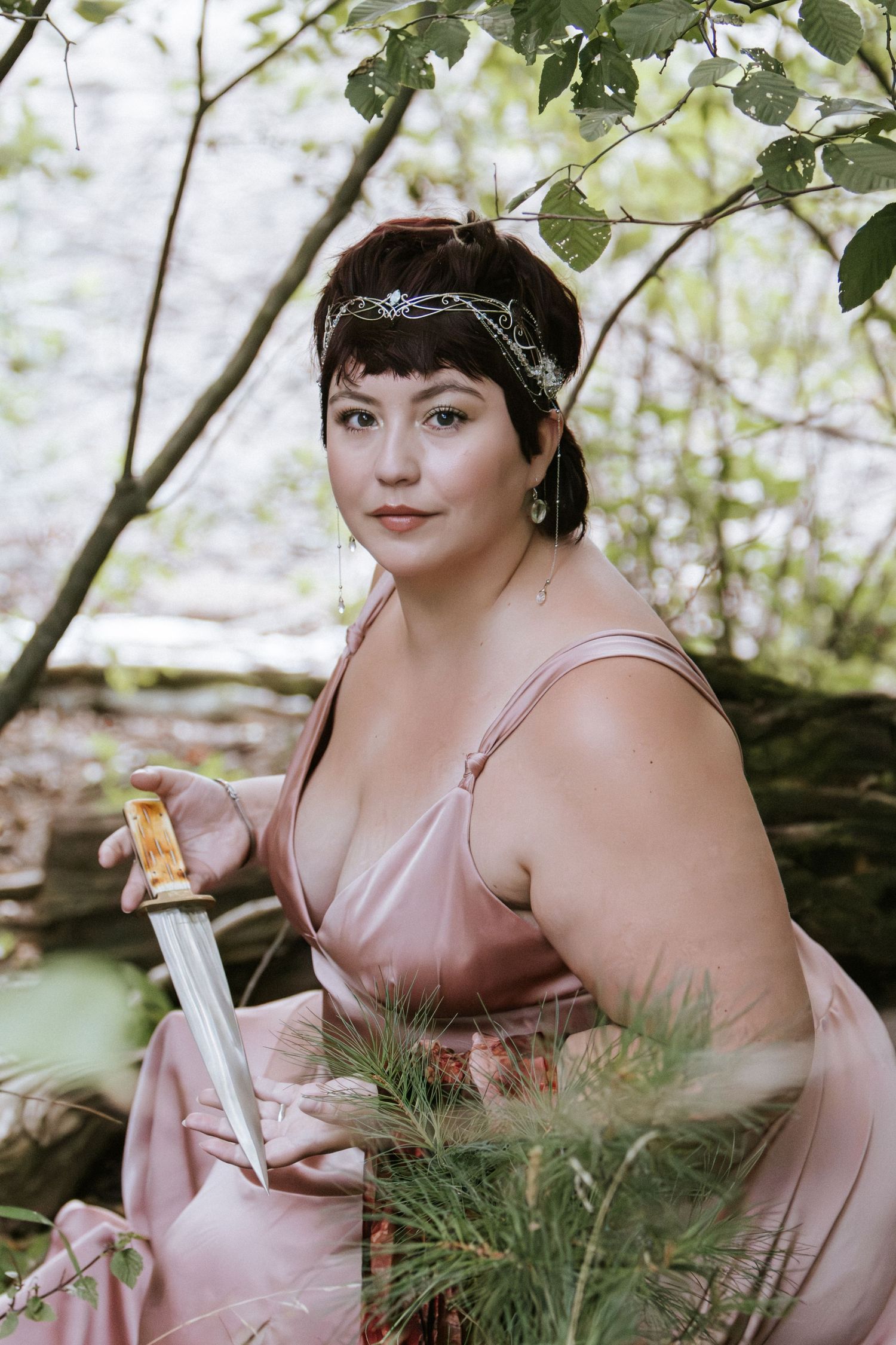 A woman in a pink satin dress and silver headpiece holds a dagger while seated among trees in a fantasy-style portrait.