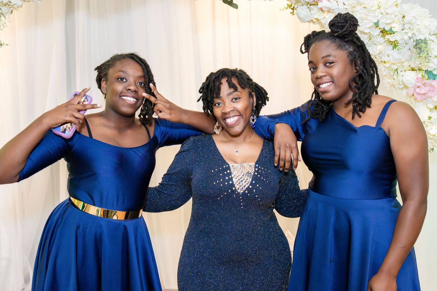 Three smiling women in royal blue dresses pose playfully together in front of a draped backdrop with white floral decor.