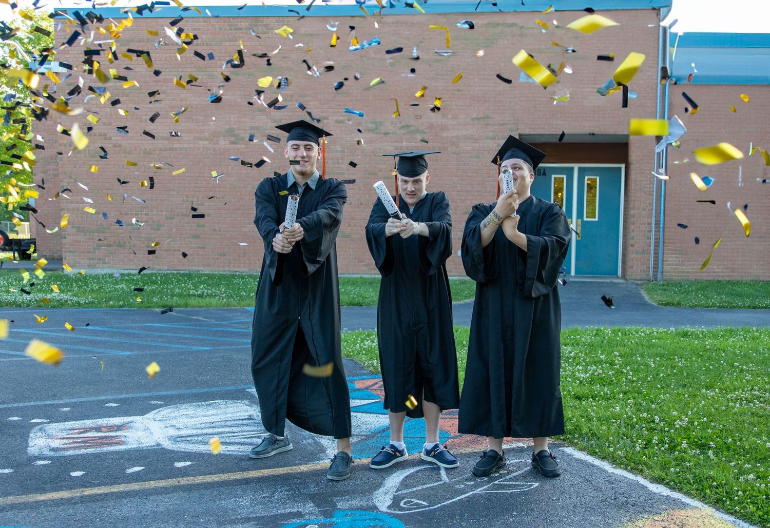 Three graduates in black caps and gowns celebrate outside a brick school building as gold and blue confetti falls around them.