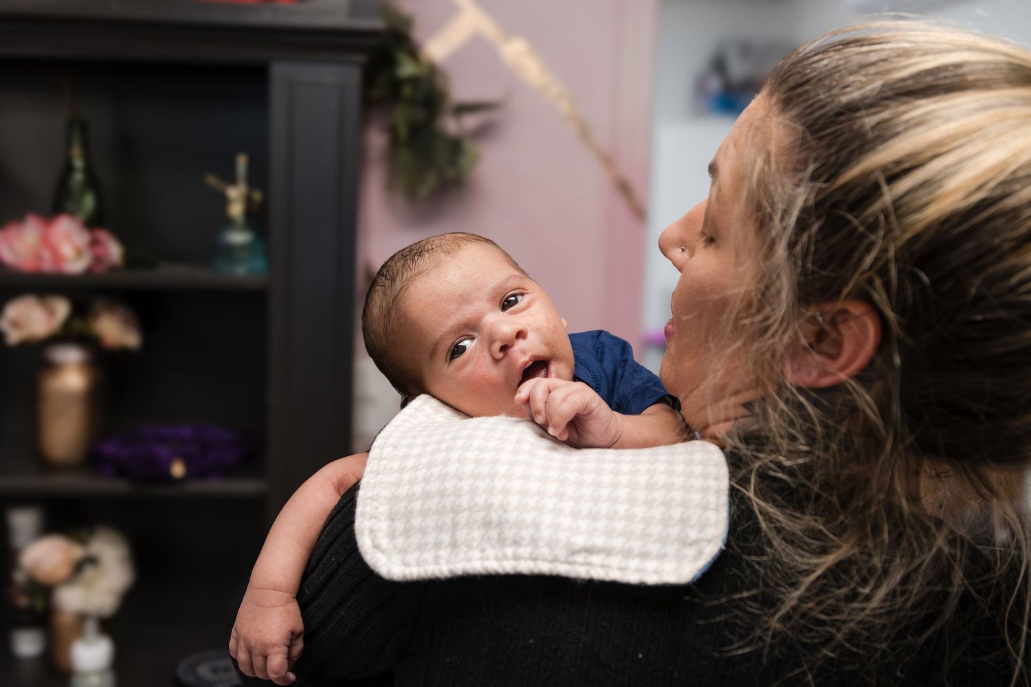 A mother holds a wide-eyed newborn baby over her shoulder with a burp cloth, in a softly lit room with decor behind them.