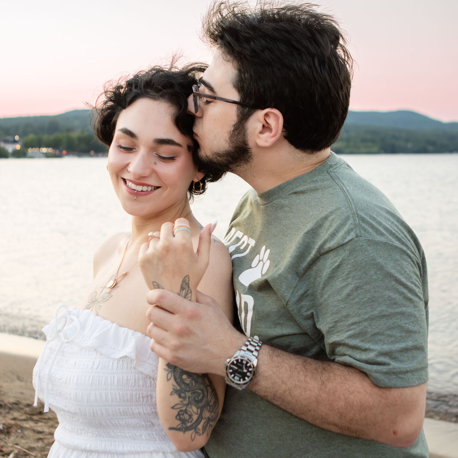 Bearded man with glasses kisses the head of a smiling woman in a white dress beside a calm lake at sunset.