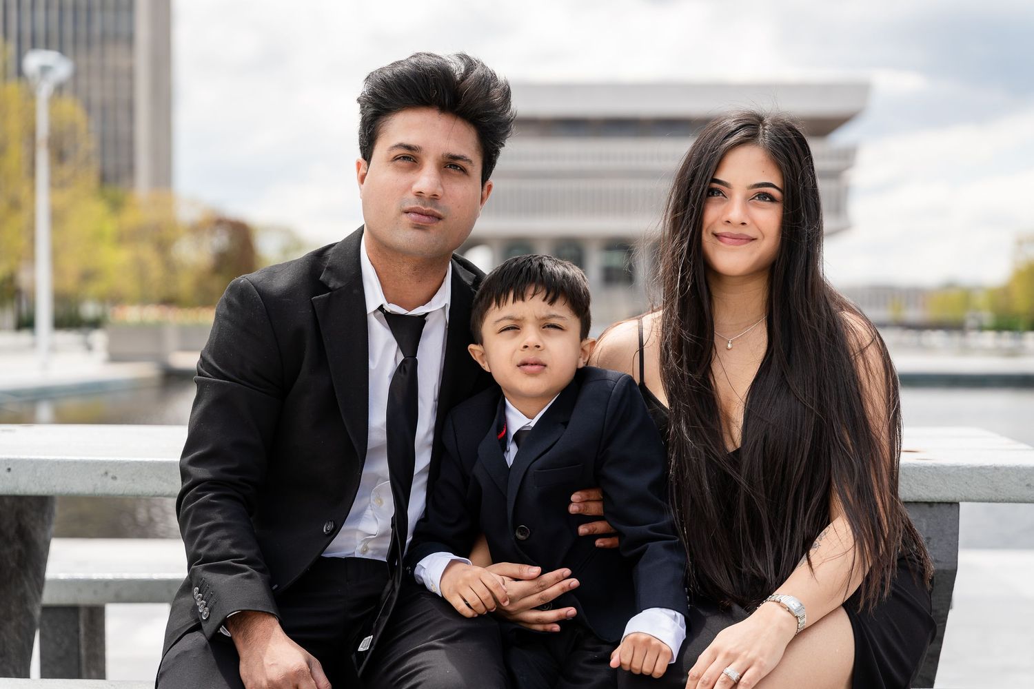 A smiling couple in black formal wear sit on a bench with their young son in a suit, with a city plaza behind them.