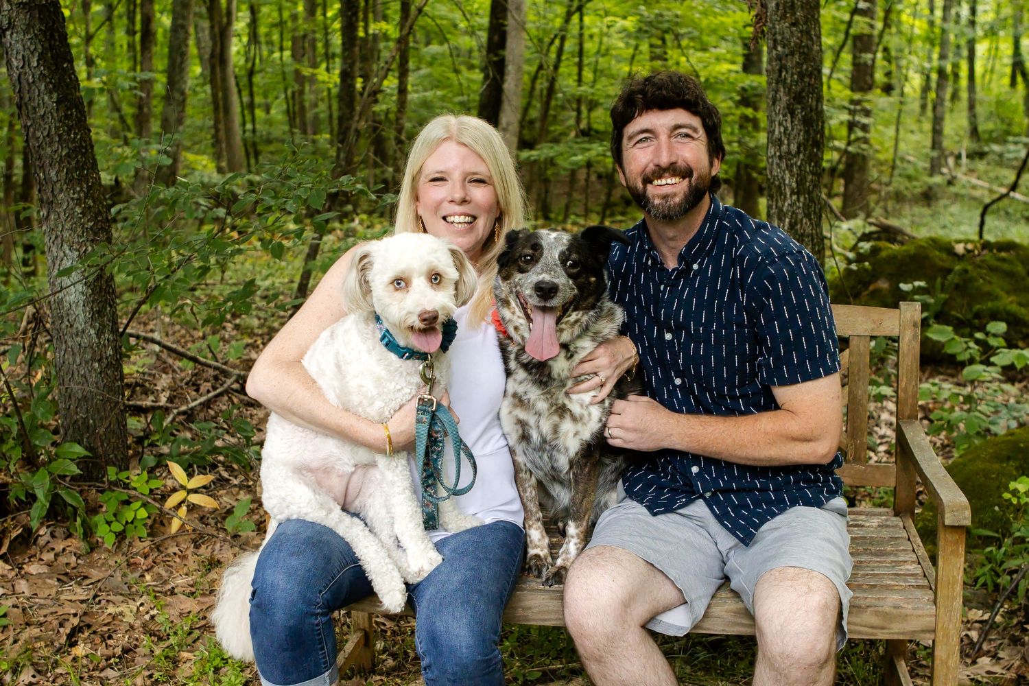 A smiling couple sits on a wooden bench in a sunlit forest, each holding one of their two dogs for a family portrait.