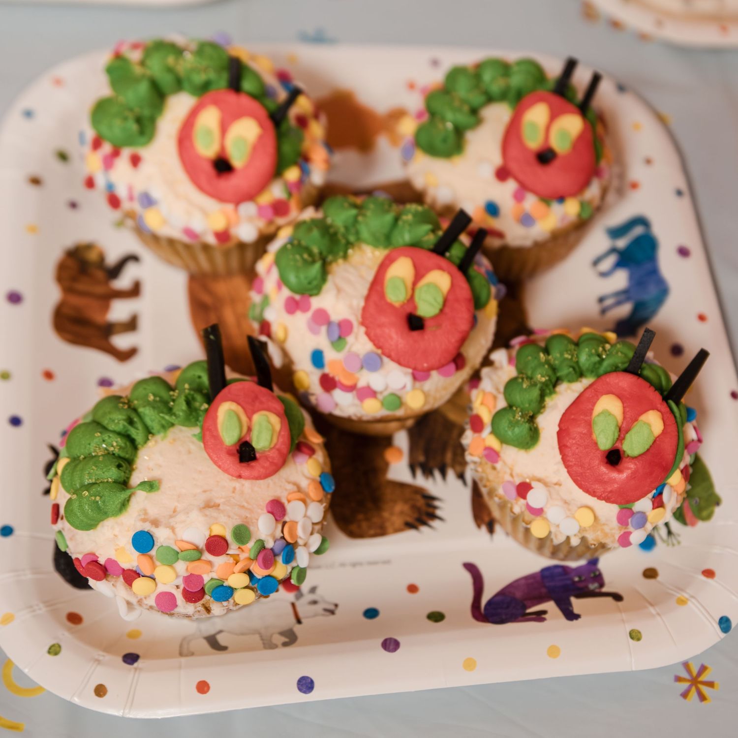Four Very Hungry Caterpillar cupcakes with green frosting bodies, red faces and sprinkles on a polka dot party plate.