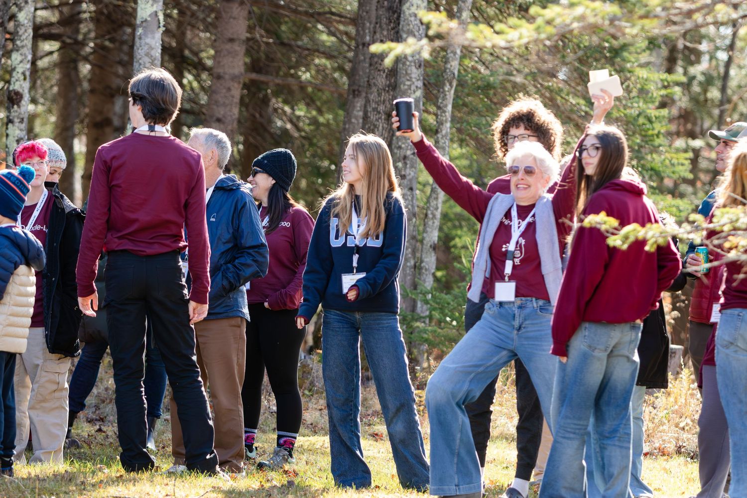 Group of people in casual jackets and jeans gathered outdoors in a sunny forest clearing, smiling while holding up phones and branches.