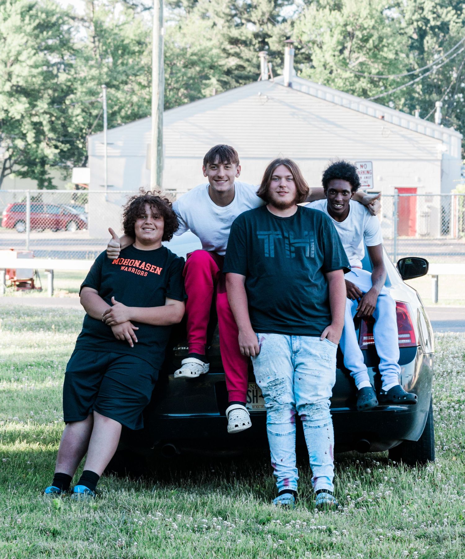 Four teenage boys pose together outdoors on the back of a black car parked on a grassy lawn on a sunny day.