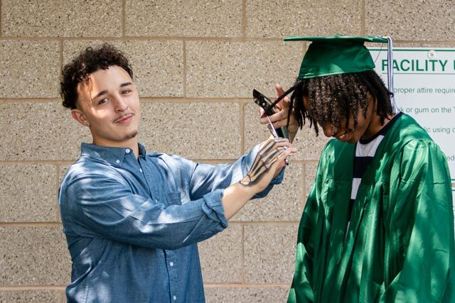 A barber with a skeleton hand tattoo trims the hair of a graduate in a green cap and gown outside a brick building.