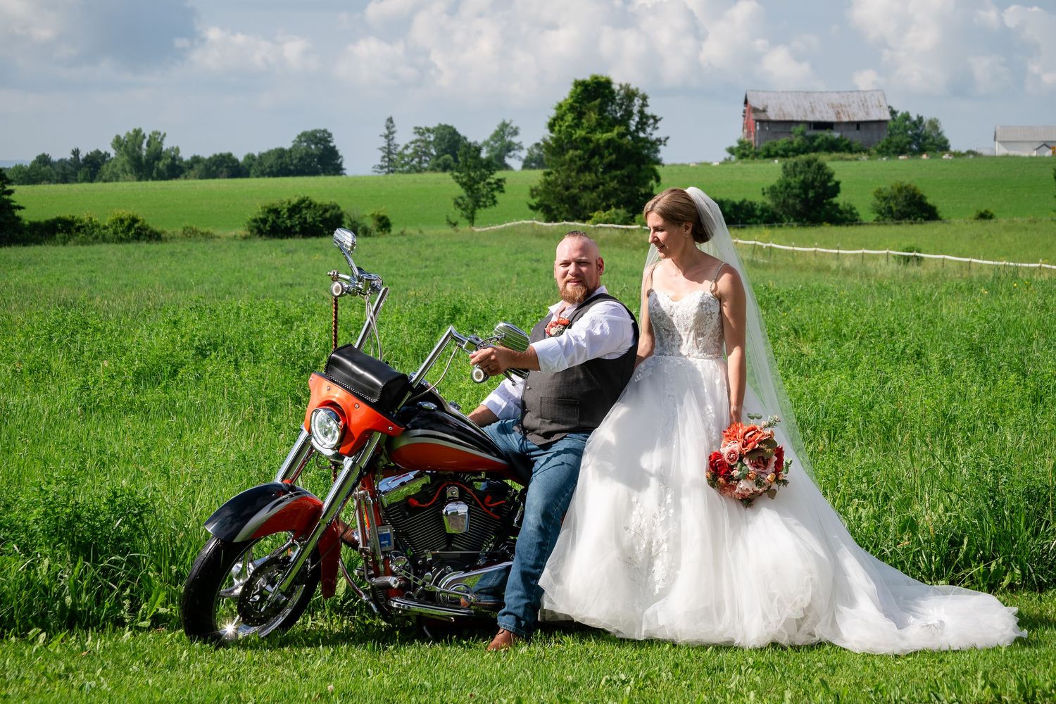 Bride in a long white gown and groom in a vest pose beside an orange motorcycle in a green farm field with a red barn in the distance.