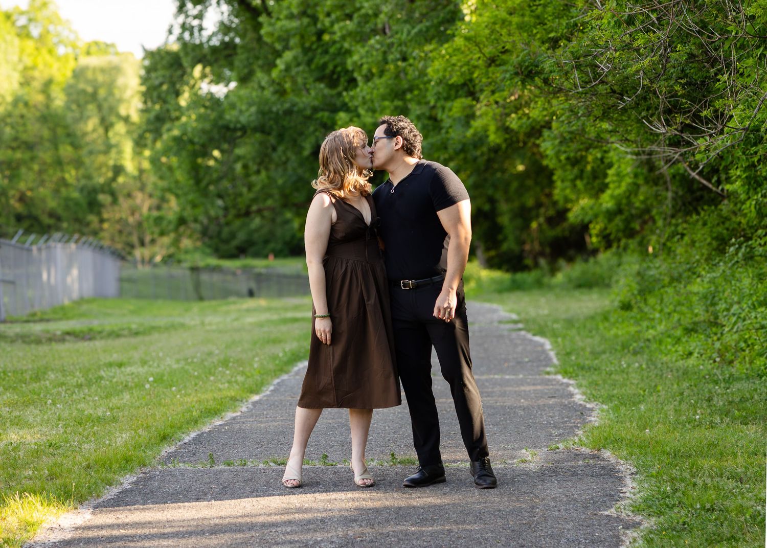 Couple kissing on a paved garden path lined with green grass and trees during an outdoor engagement photo session.